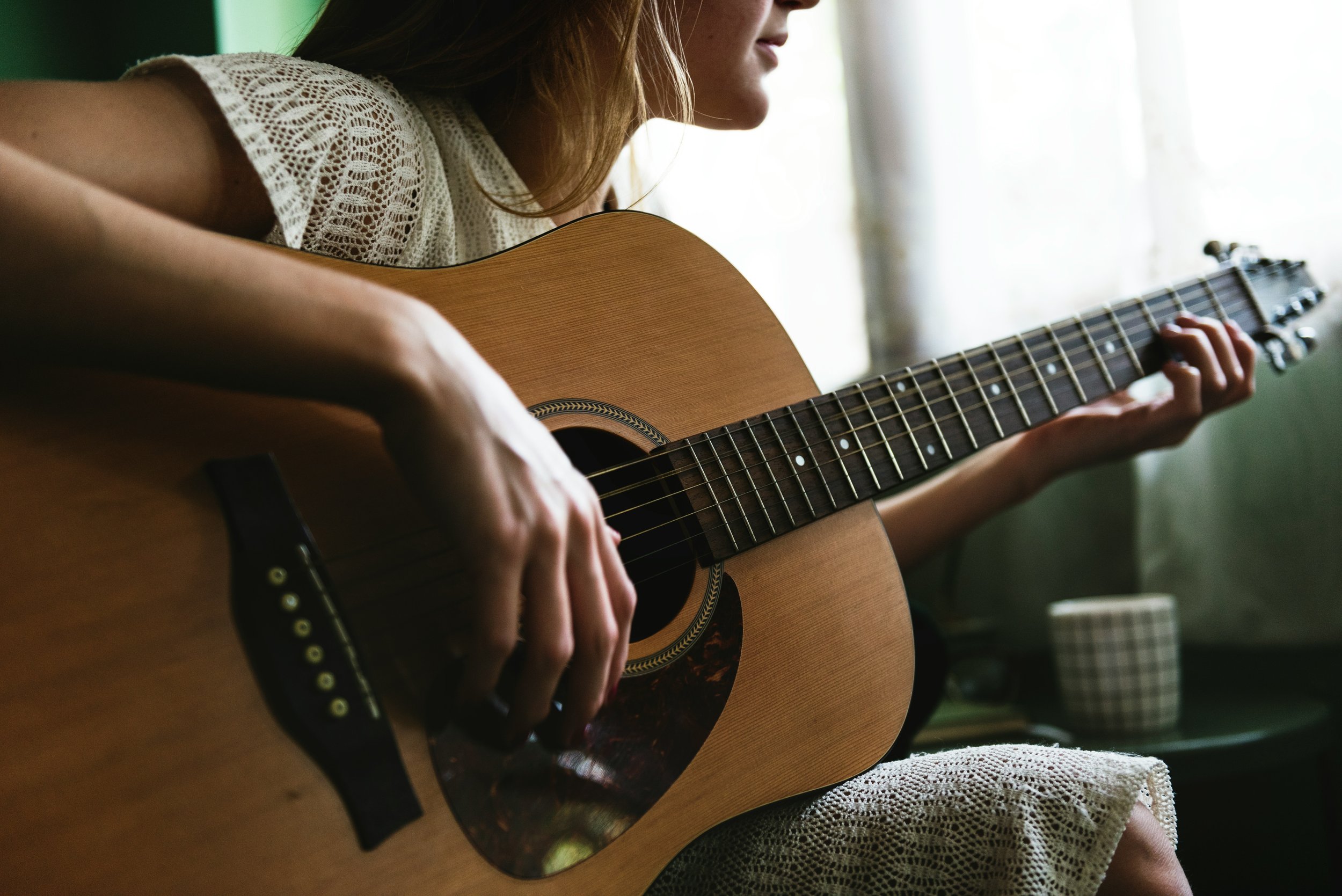 Woman playing an acoustic guitar indoors near a window with natural light.