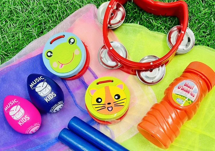 A collection of colorful children's toys on a multicolored fabric surface, including a frog face button, a tiger face button, a red tambourine, a neon orange juice bottle, and two blue swim noodles.