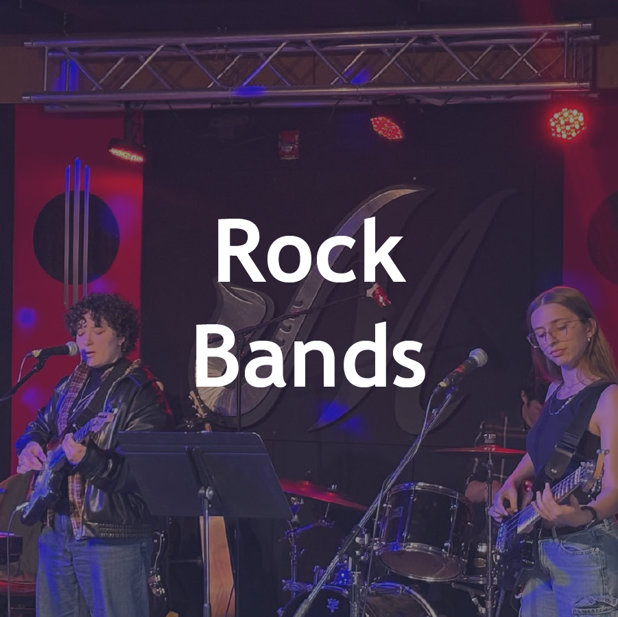 Two young women playing electric guitars and singing into microphones on stage with a drum set in the background, under red and blue stage lights at a live music venue.