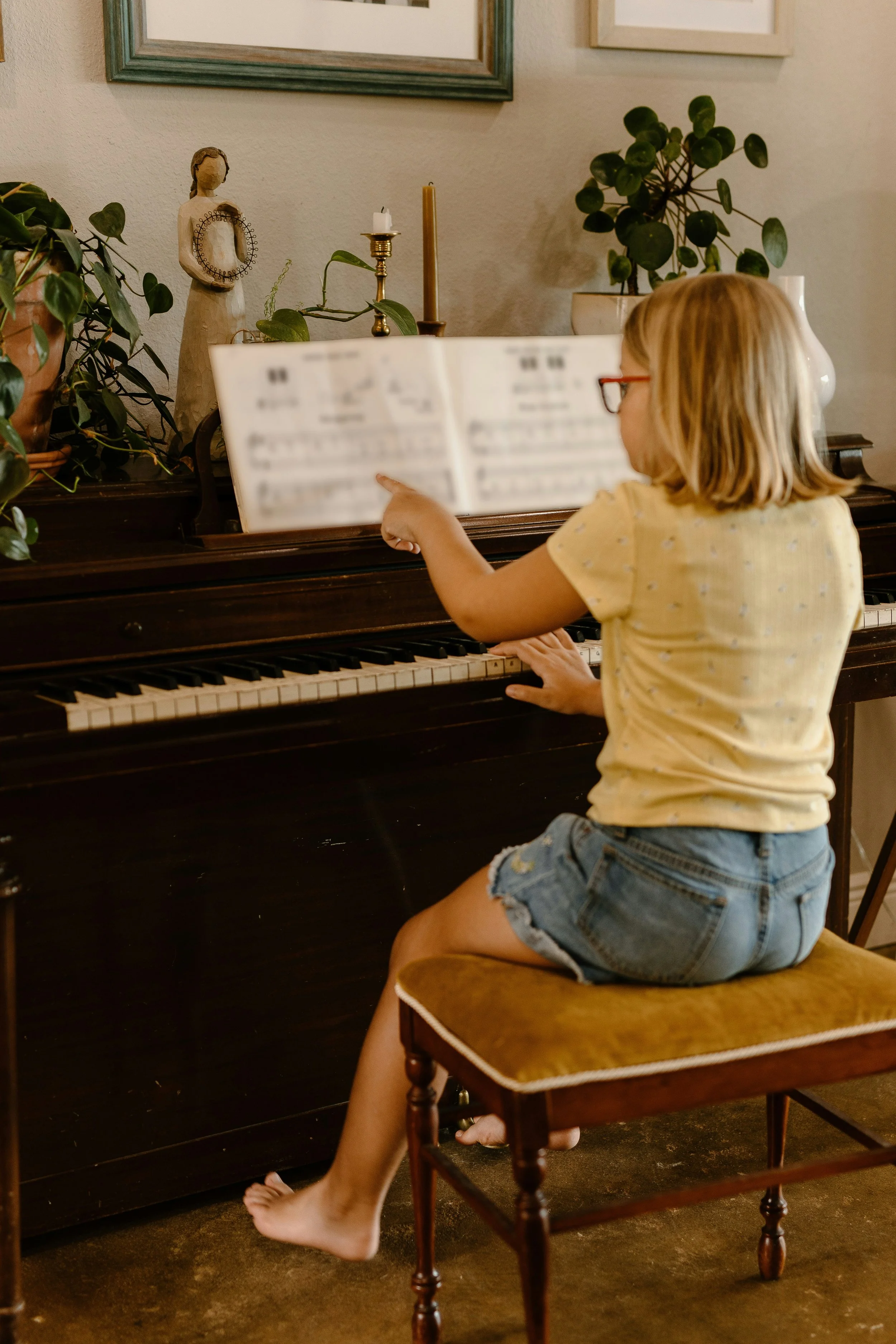 A young girl with blonde hair, glasses, a yellow shirt, and denim shorts sitting on a yellow cushioned wooden chair, pointing at music notes on a sheet music book while sitting at a dark wooden upright piano in a home setting with houseplants and framed pictures on the wall.