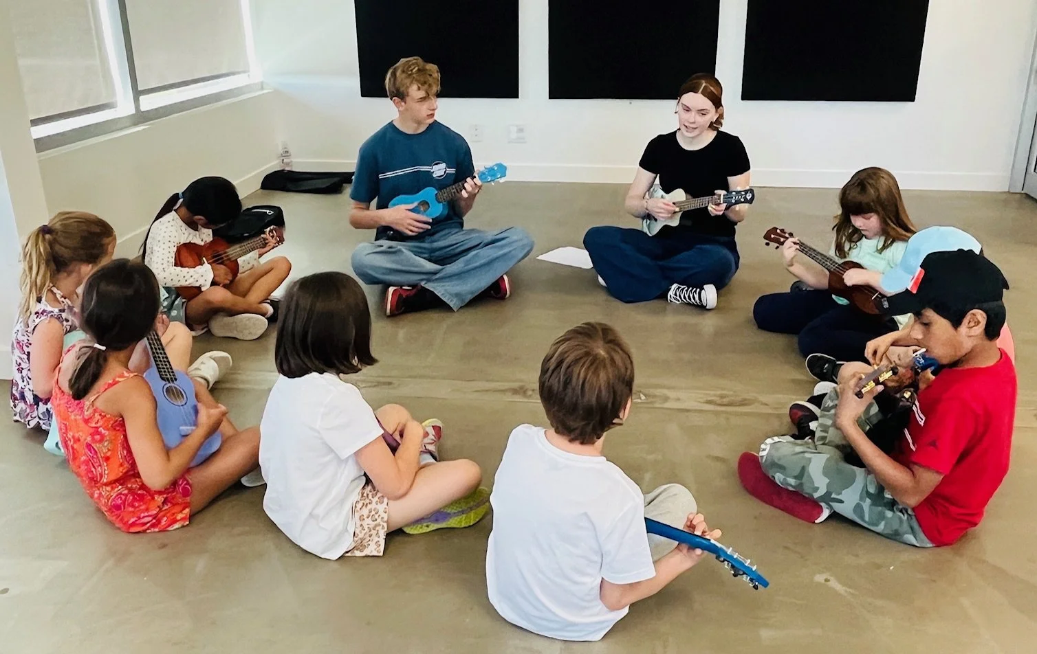 Children and two adults sitting in a circle, playing ukuleles indoors.