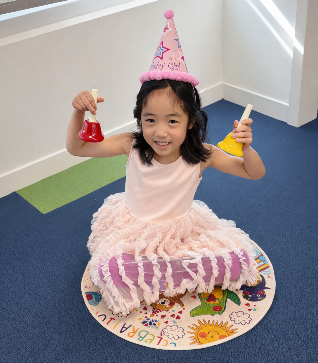 A young girl wearing a pink birthday hat and a pink dress with a ruffled skirt, sitting on a colorful circular rug with birthday illustrations, holding small party bells, celebrating her birthday.
