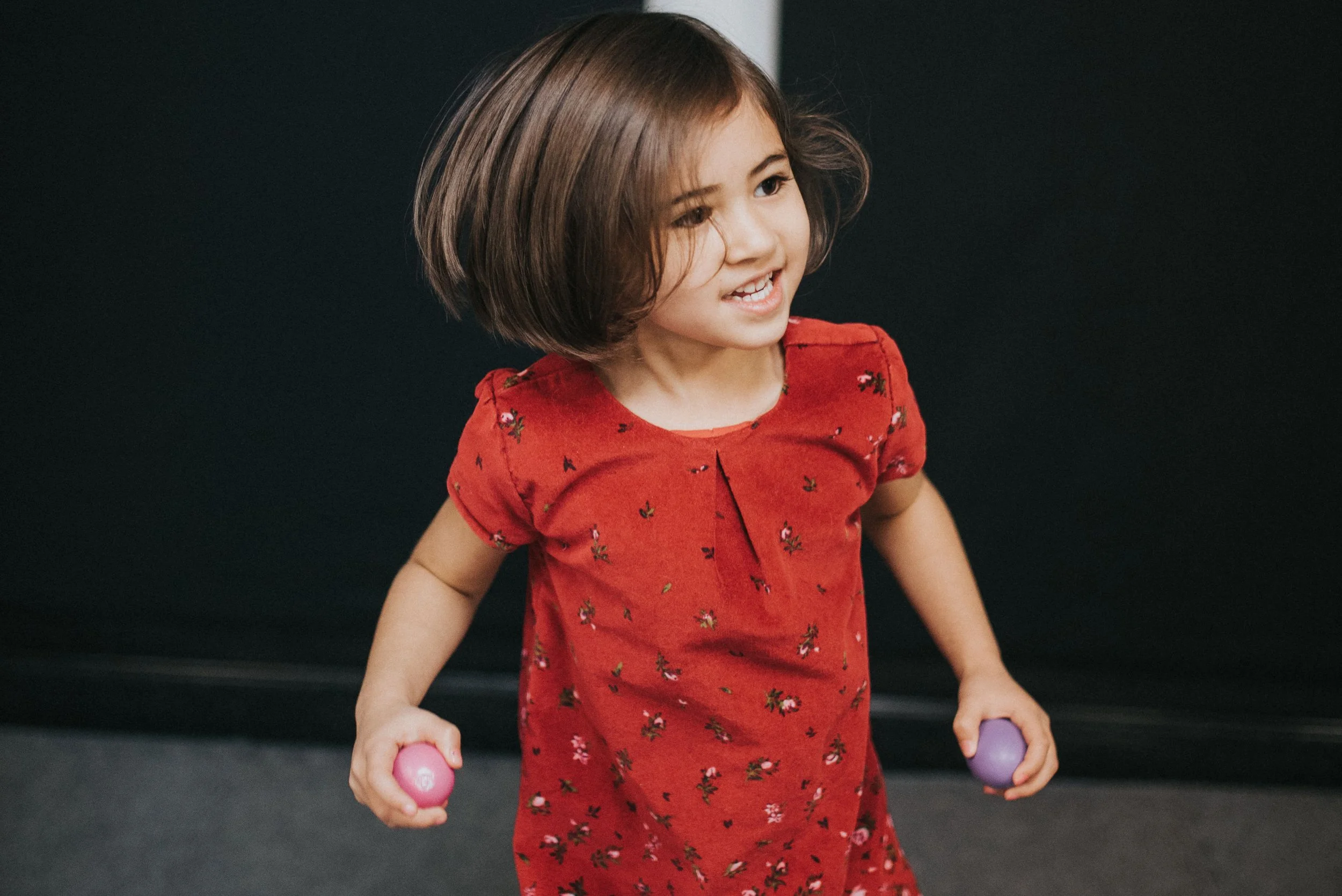 A young girl with short brown hair smiling, holding a pink ball in her right hand and a purple ball in her left hand, wearing a red dress with small floral patterns, standing against a dark background.