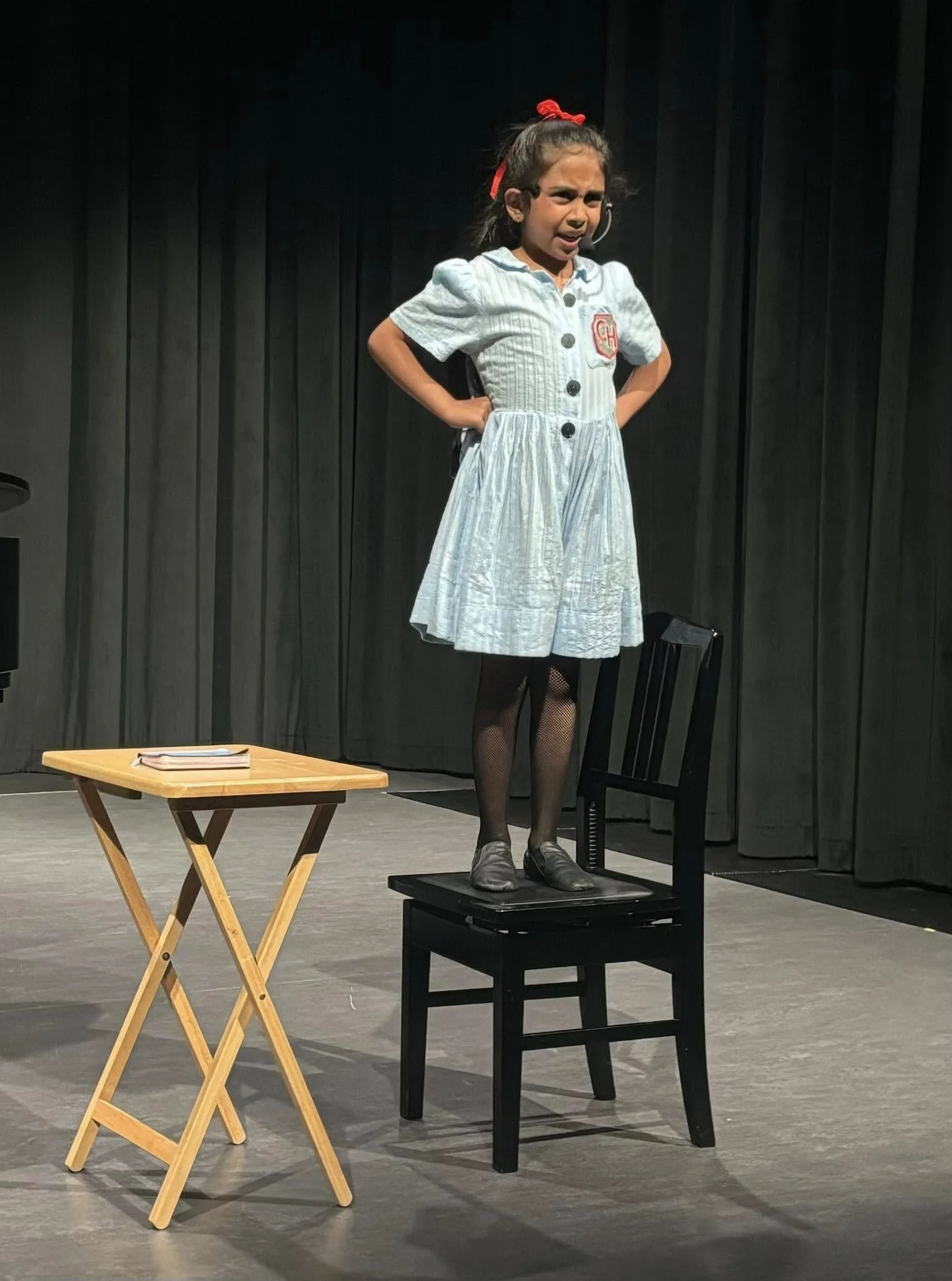 Young girl on stage, standing on a chair, wearing a light blue dress with black tights and shoes, with hands on her hips, performing with a serious expression, in front of black curtains.
