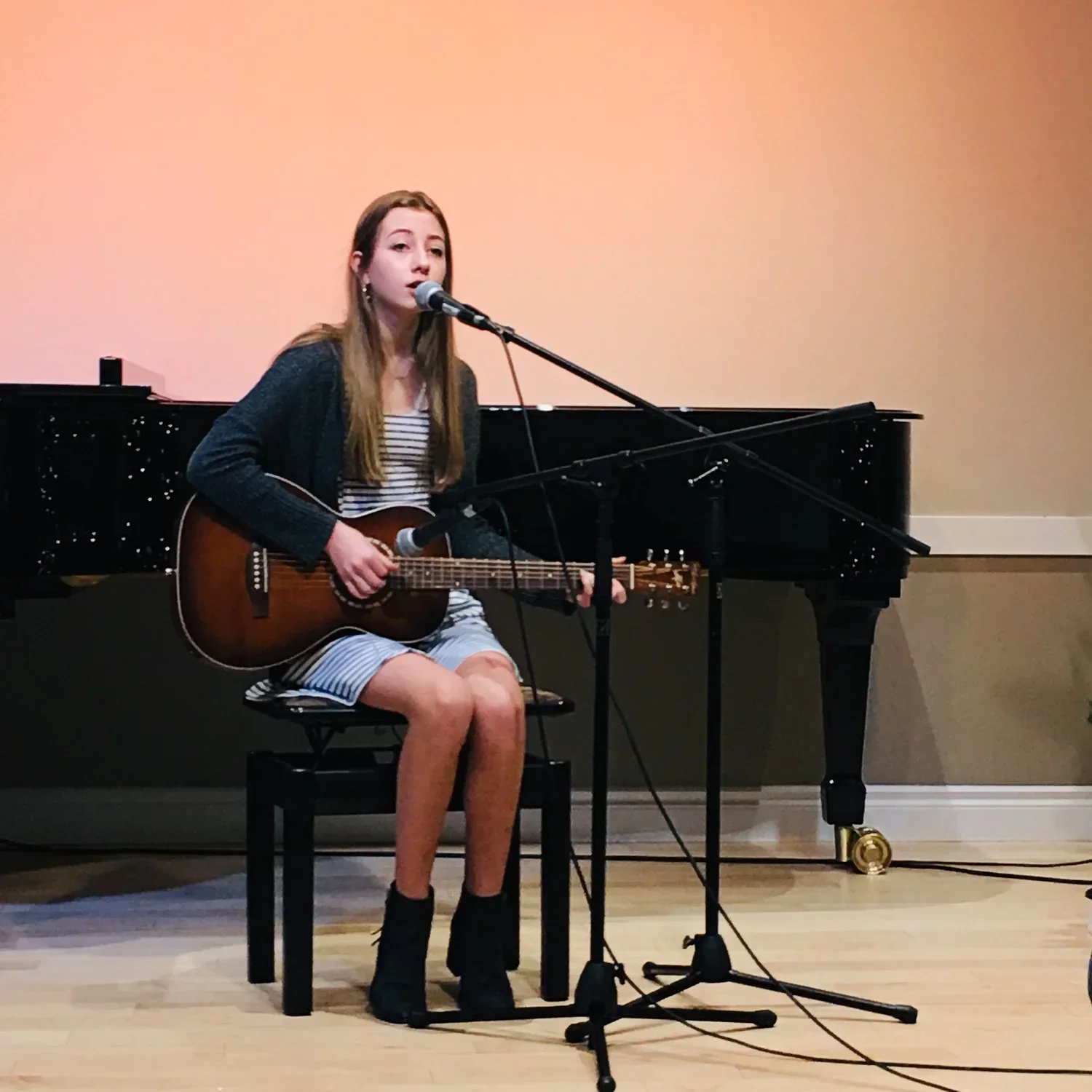 A young woman with long brown hair sitting on a black bench, singing into a microphone and playing an acoustic guitar during a performance, with a grand piano behind her.