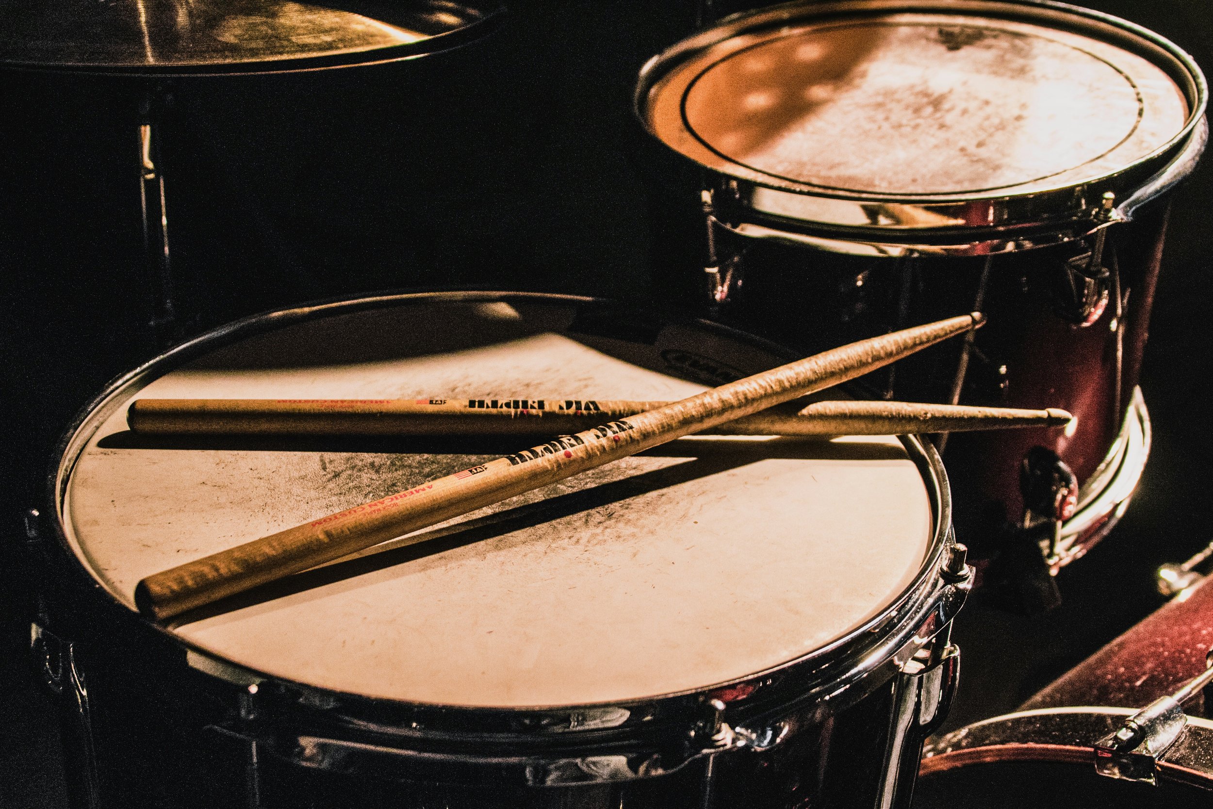 Close-up of two drums with drumsticks resting on the drumhead of the front drum, both drums are black with shiny metal rims, in a dimly lit setting.