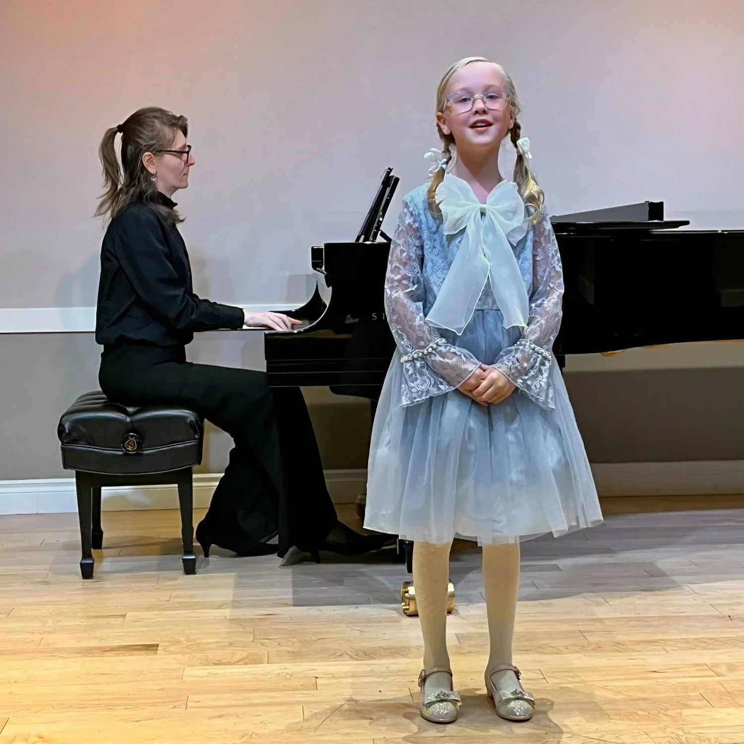 A young girl in a blue dress with a large bow on the front, standing in front of a black grand piano, with an older woman playing piano in the background.