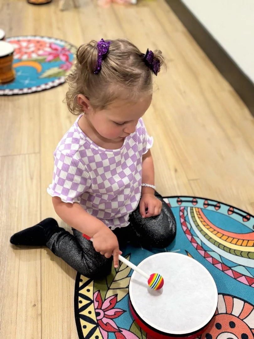 A young girl sitting on a colorful, patterned rug, playing with a white drum and a striped ball on top of it.