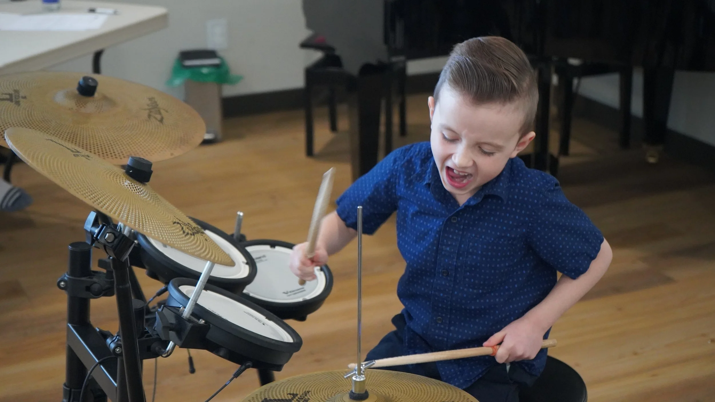 A young boy with short, neatly styled hair wearing a blue button-up shirt playing an electronic drum set passionately.
