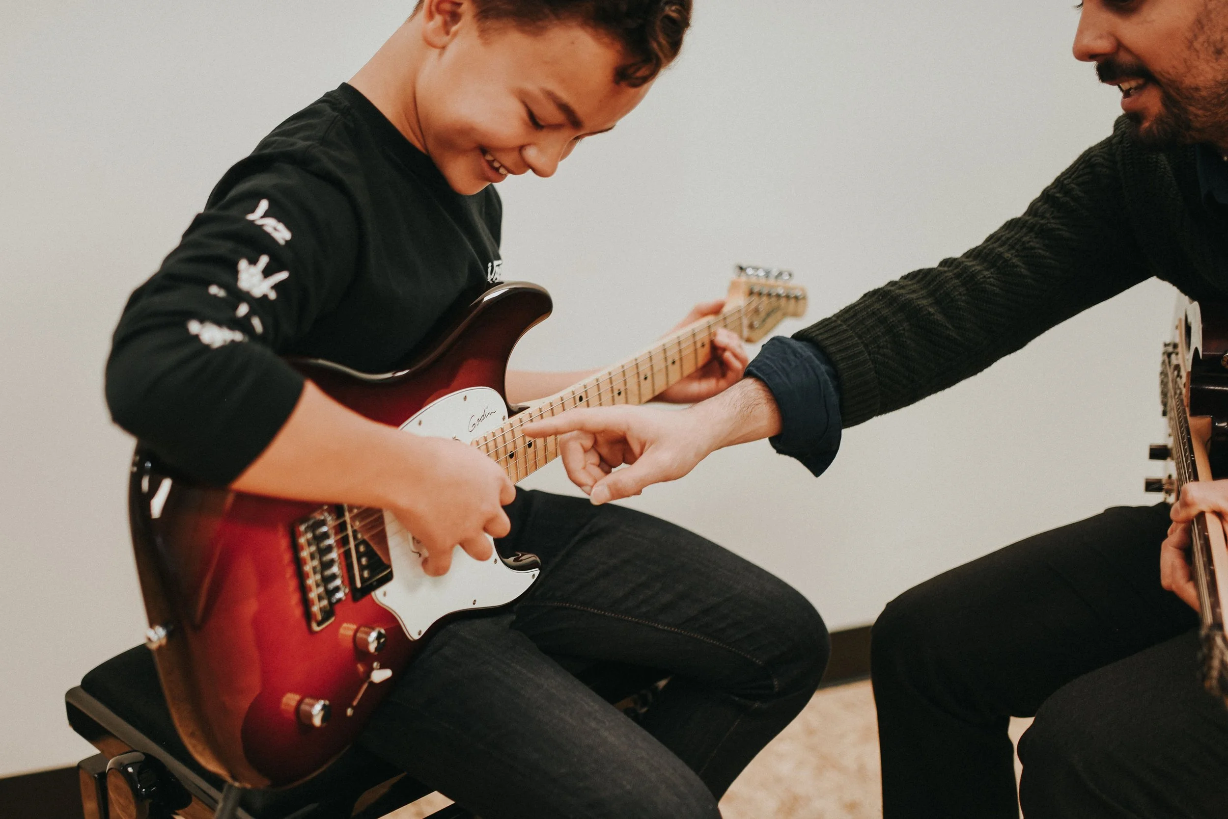 A young person and an older person are sitting together, playing guitars. The younger person is smiling and pointing at the guitar while the older person, who is holding another guitar, looks on. They are in a room with plain white walls.