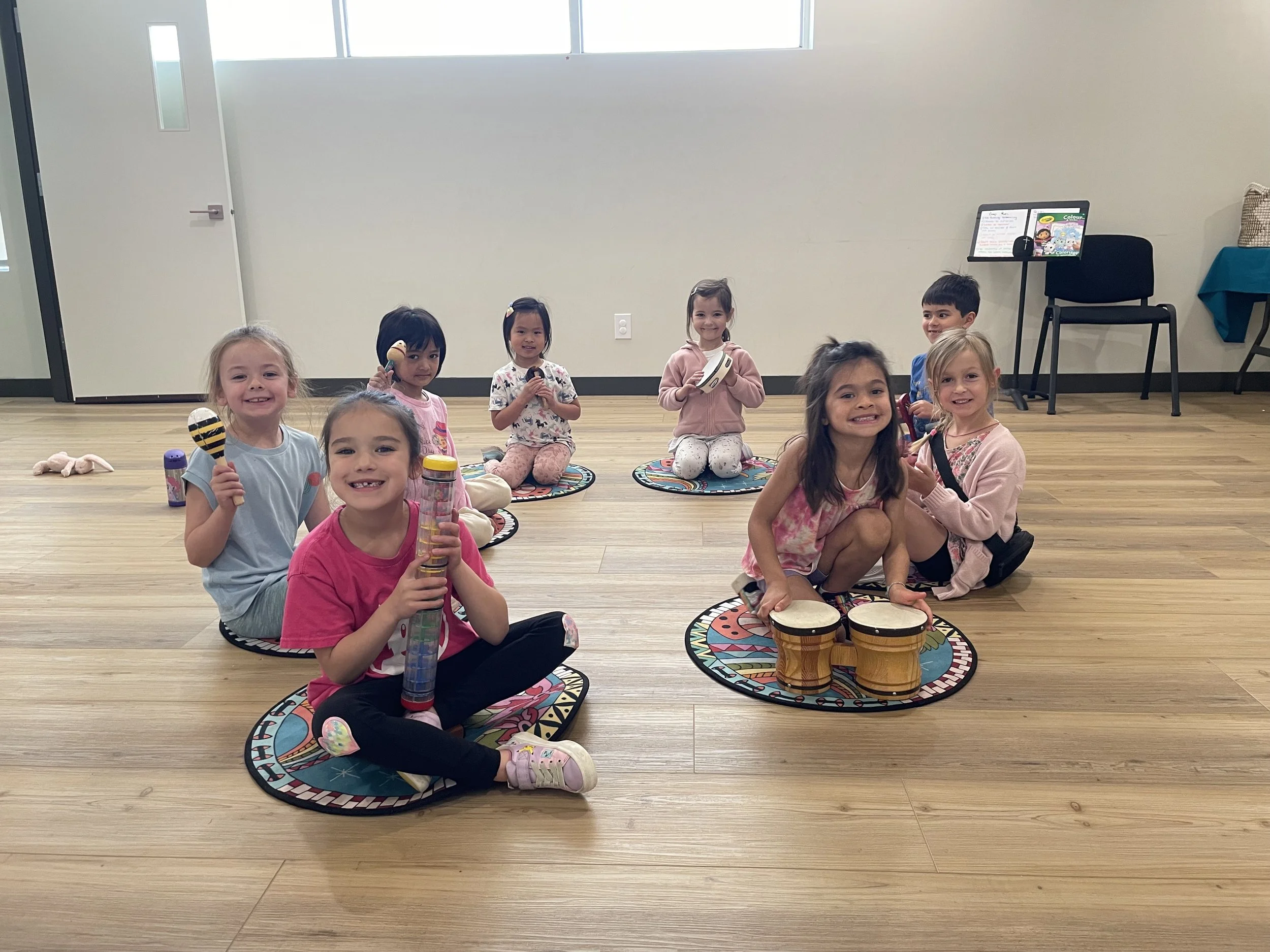 Group of children sitting on colorful circular mats, holding musical instruments like maracas, tambourines, and drums, smiling in a classroom with wooden floors and white walls.