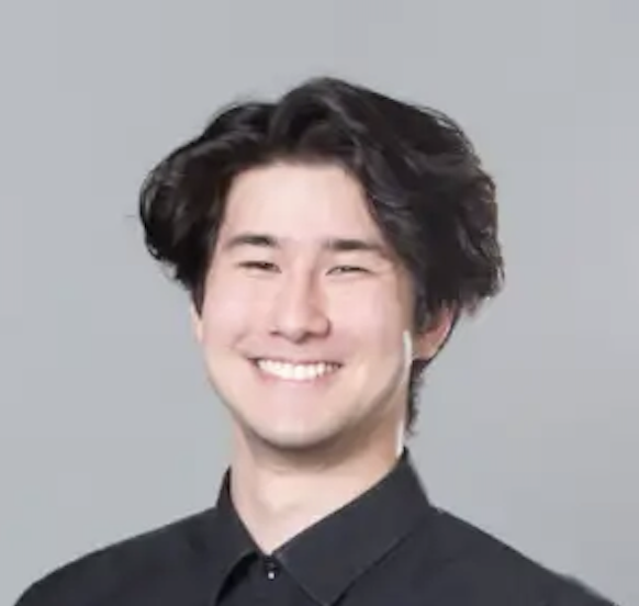 Smiling young man with dark, curly hair and wearing a black shirt against a gray background.