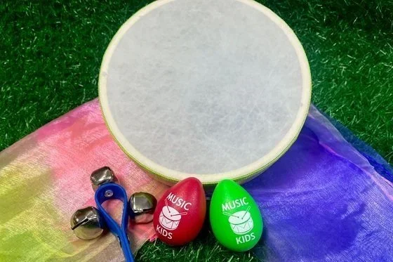 A circular drum surface, a pair of colorful egg-shaped percussion instruments labeled "Music Kids," a pair of small metal bells, and a pair of rainbow-colored fabric pieces are arranged on artificial grass.