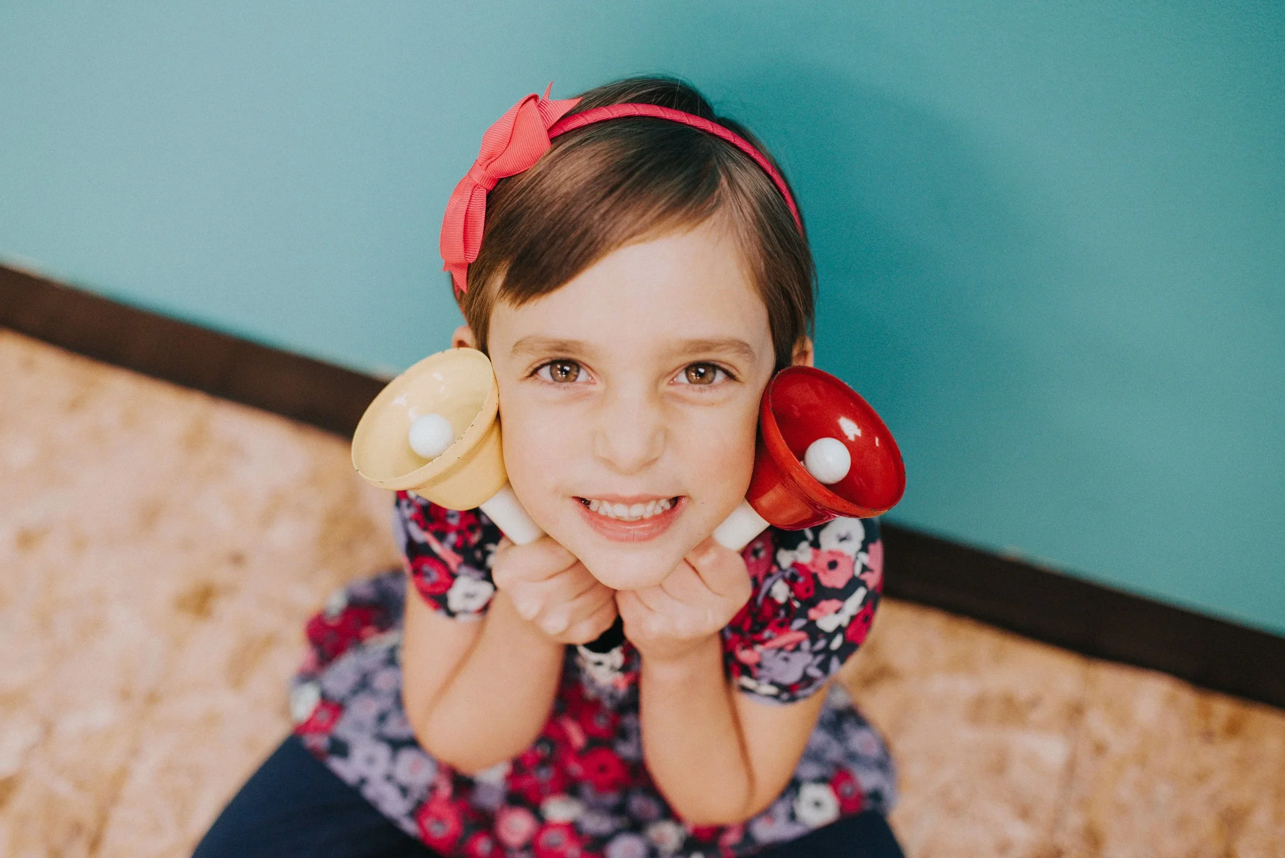 A young girl with a pink bow headband sitting on the floor, smiling, with toy bowls on her ears and looking up at the camera.