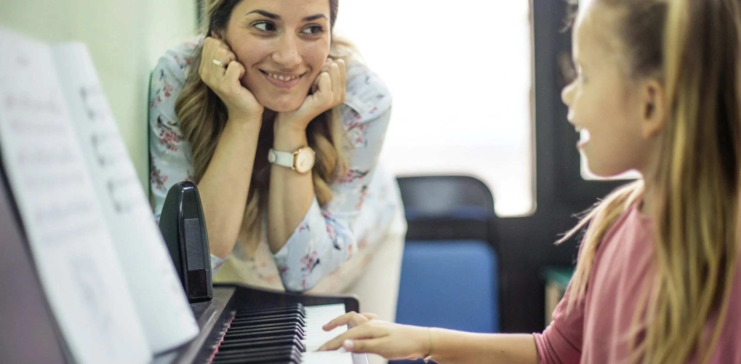 A woman and a young girl are sharing a music lesson at a piano. The woman, smiling, leans on her hands while observing the girl play the piano.