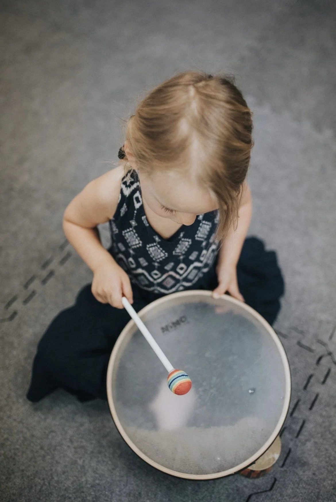 A young girl sitting cross-legged on the floor, holding a drumstick, about to strike a small round drum with a colorful striped ball attached to a stick.