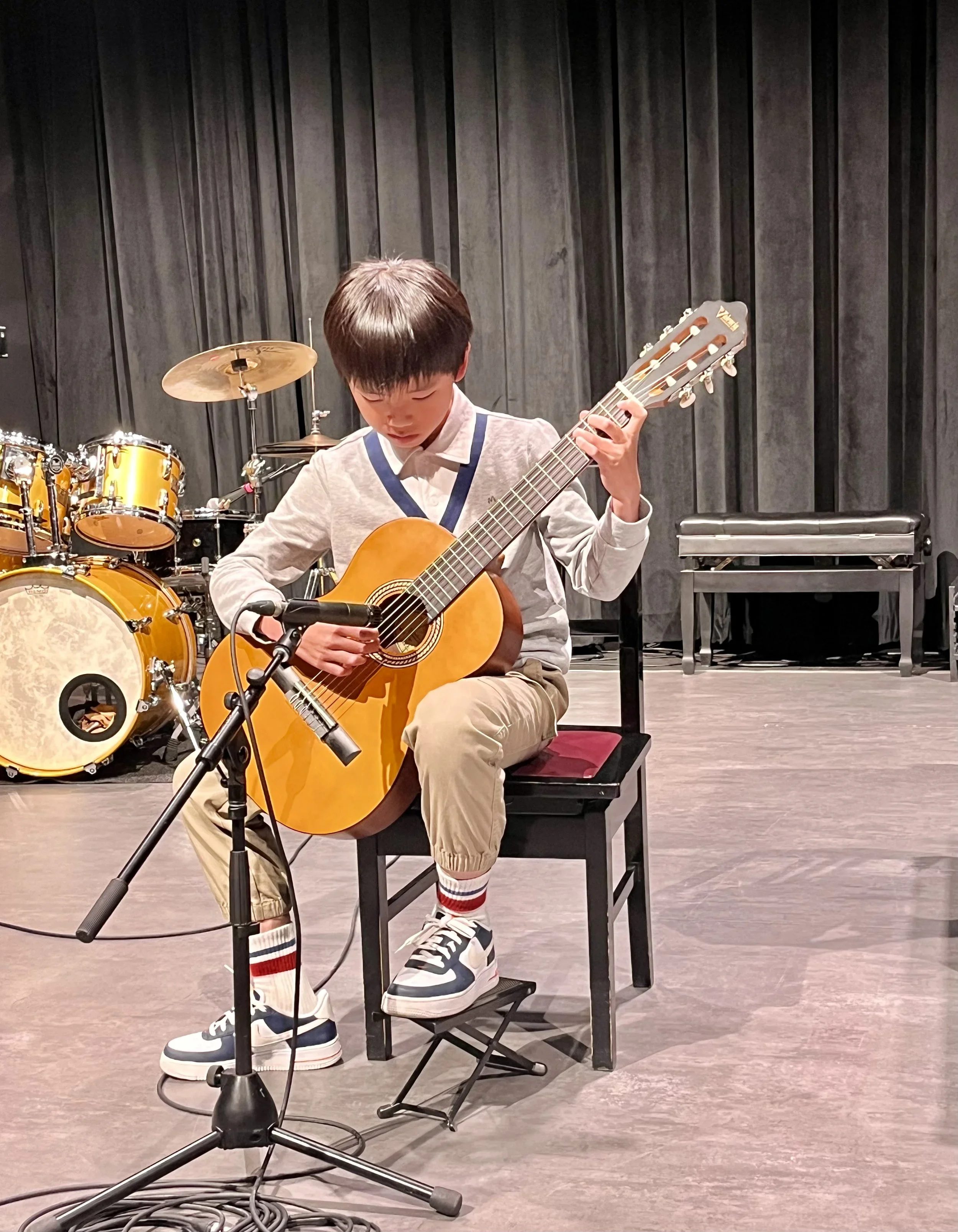 A young boy is sitting on a chair on stage, playing an acoustic guitar. There are drums in the background, and a microphone stand in front of him.