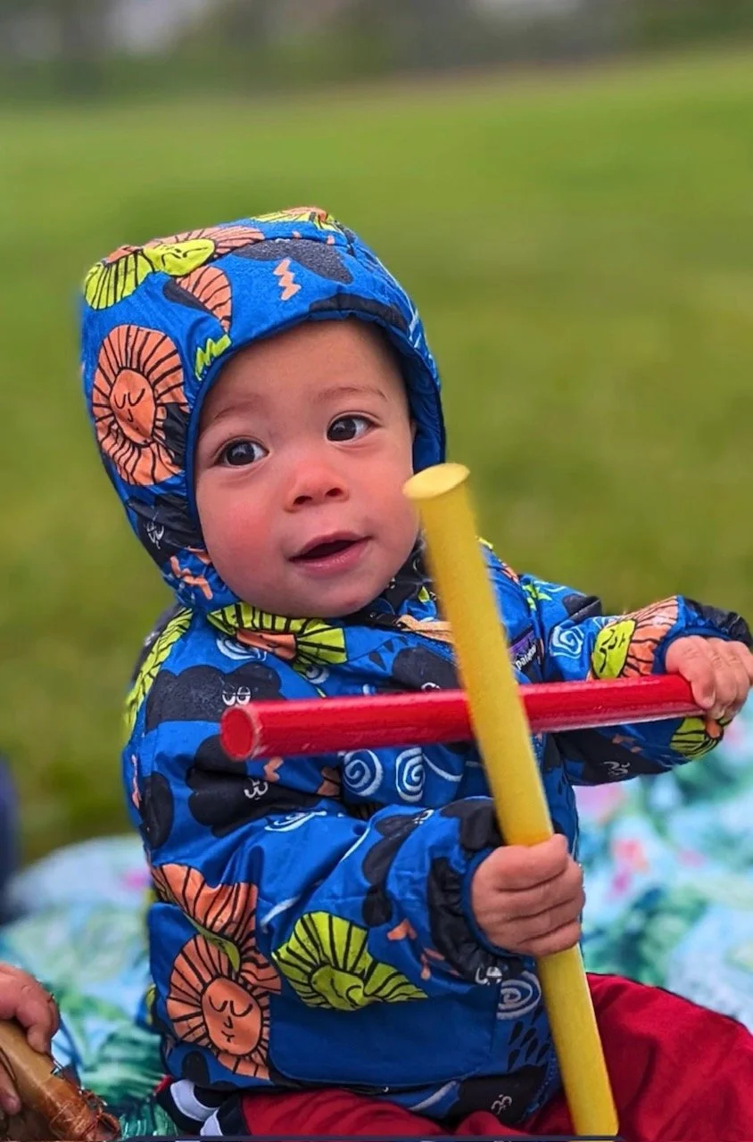 A young child wearing a colorful, patterned raincoat and hat outdoors, holding a toy with a yellow pole and red crossbar, with a grassy background.