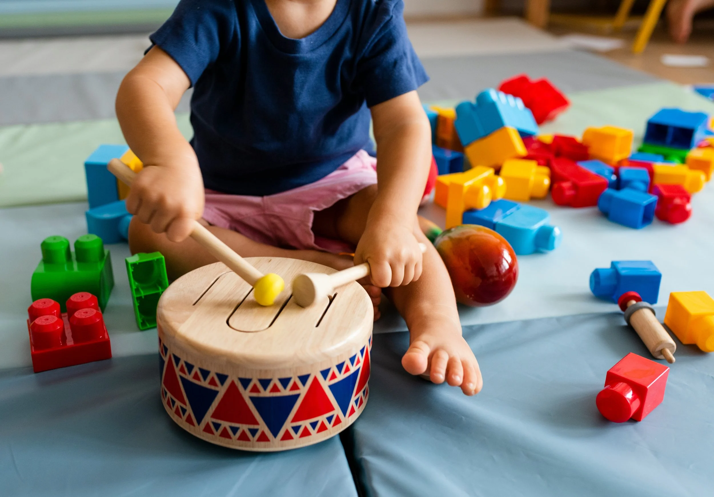 A child playing with a wooden drum and colorful plastic building blocks on a blue surface.