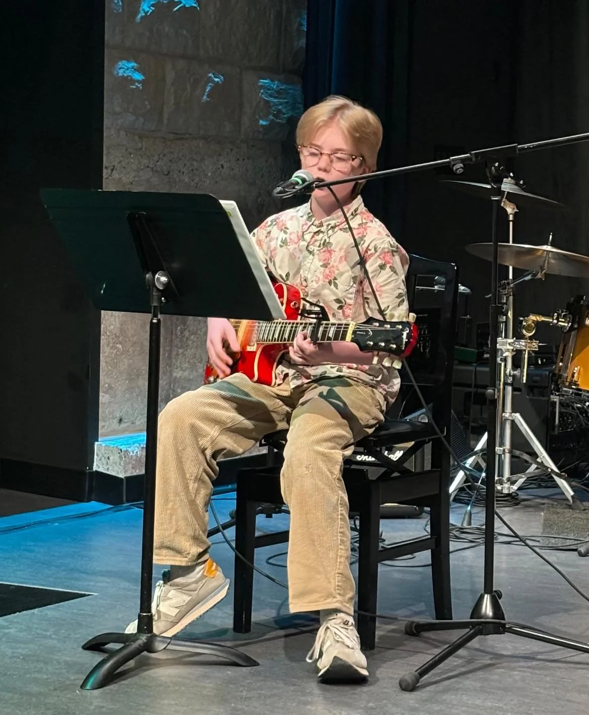 A person with glasses and a happy expression, seated on a black chair, playing a red electric guitar and singing into a microphone during a musical performance. The person is wearing a floral shirt, beige pants, and white sneakers, with music stands and drum set equipment nearby on a stage with dark background and blue lighting accents.