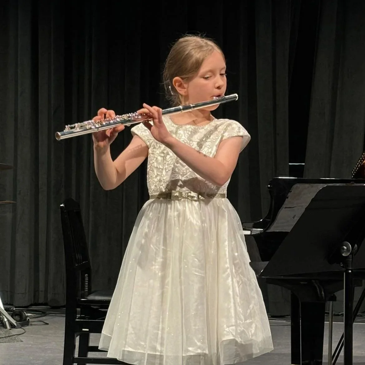 Young girl in a fancy dress playing the flute on stage with a piano and black curtains in the background.
