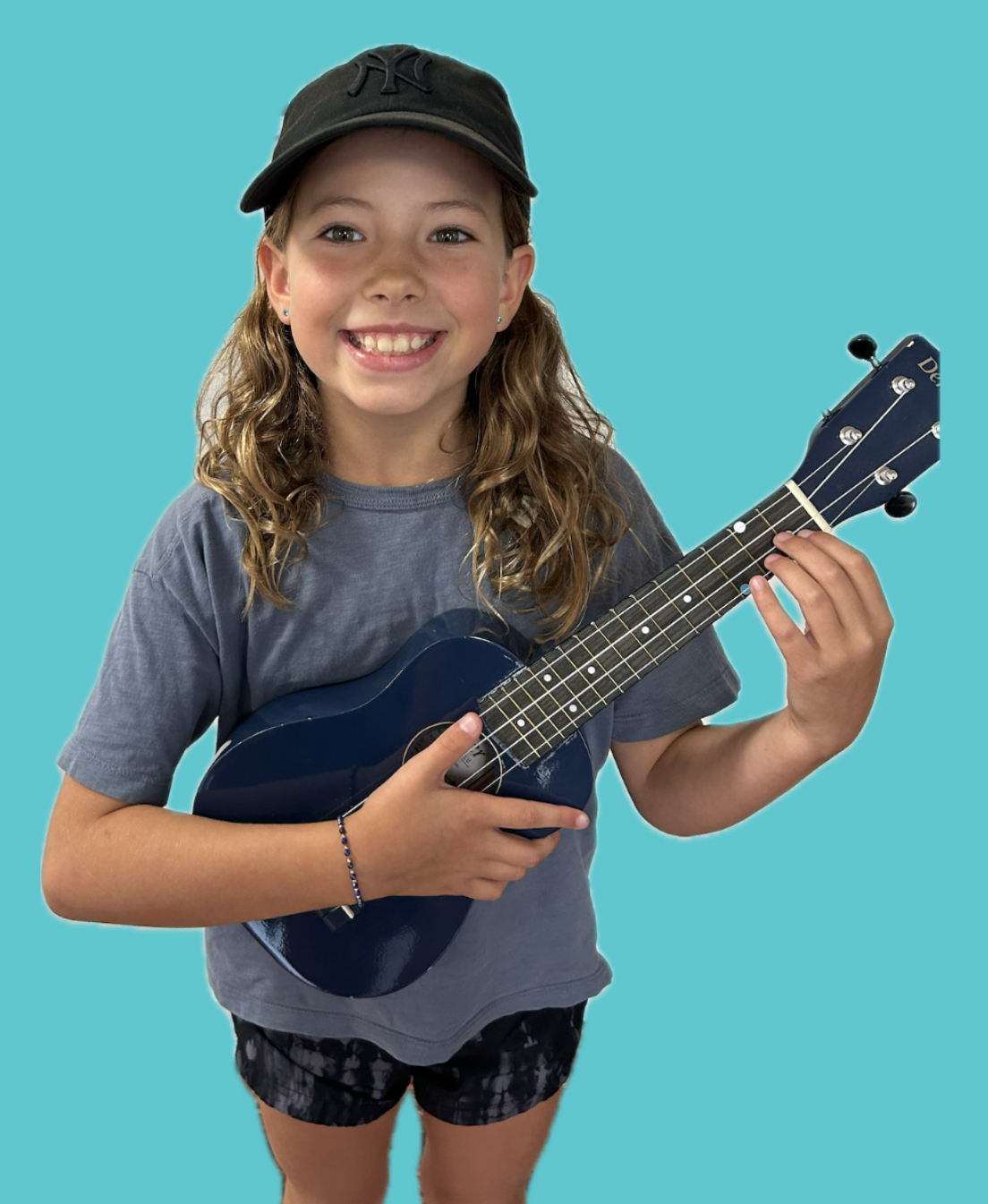 A young girl with curly hair, wearing a black cap, gray t-shirt, and patterned shorts, smiling and holding a small blue ukulele against a solid light blue background.