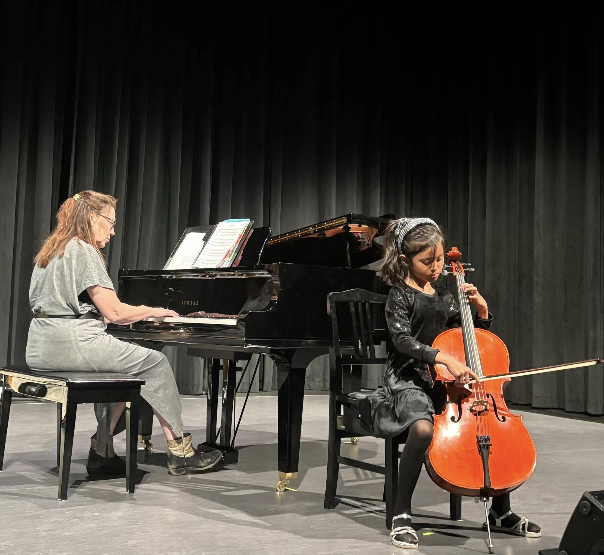 A woman playing the piano and a girl playing the cello on a stage with a black curtain background.