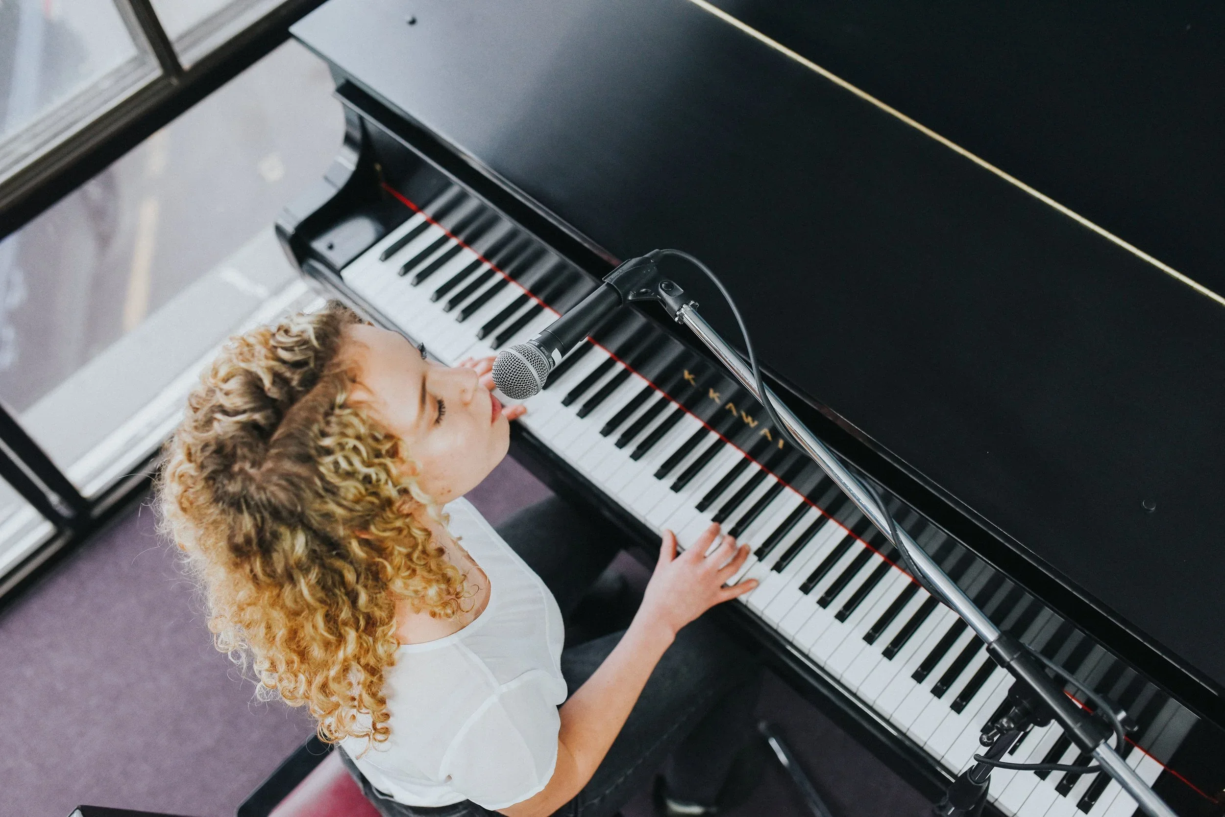 A young woman with curly blonde hair playing a grand piano, singing into a microphone.