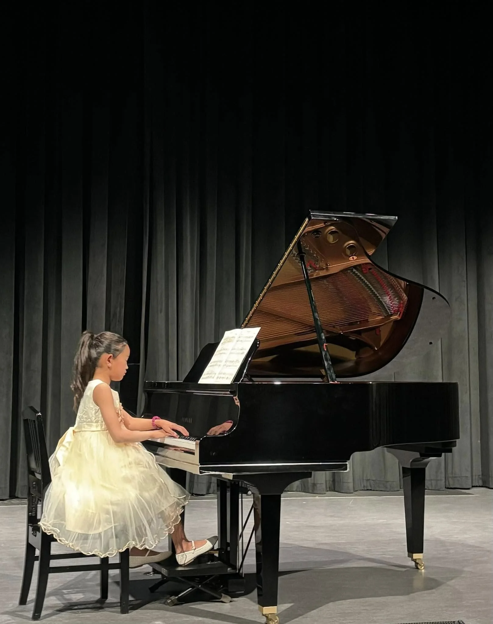 A young girl in a yellow dress playing the grand piano on stage with a black curtain backdrop.