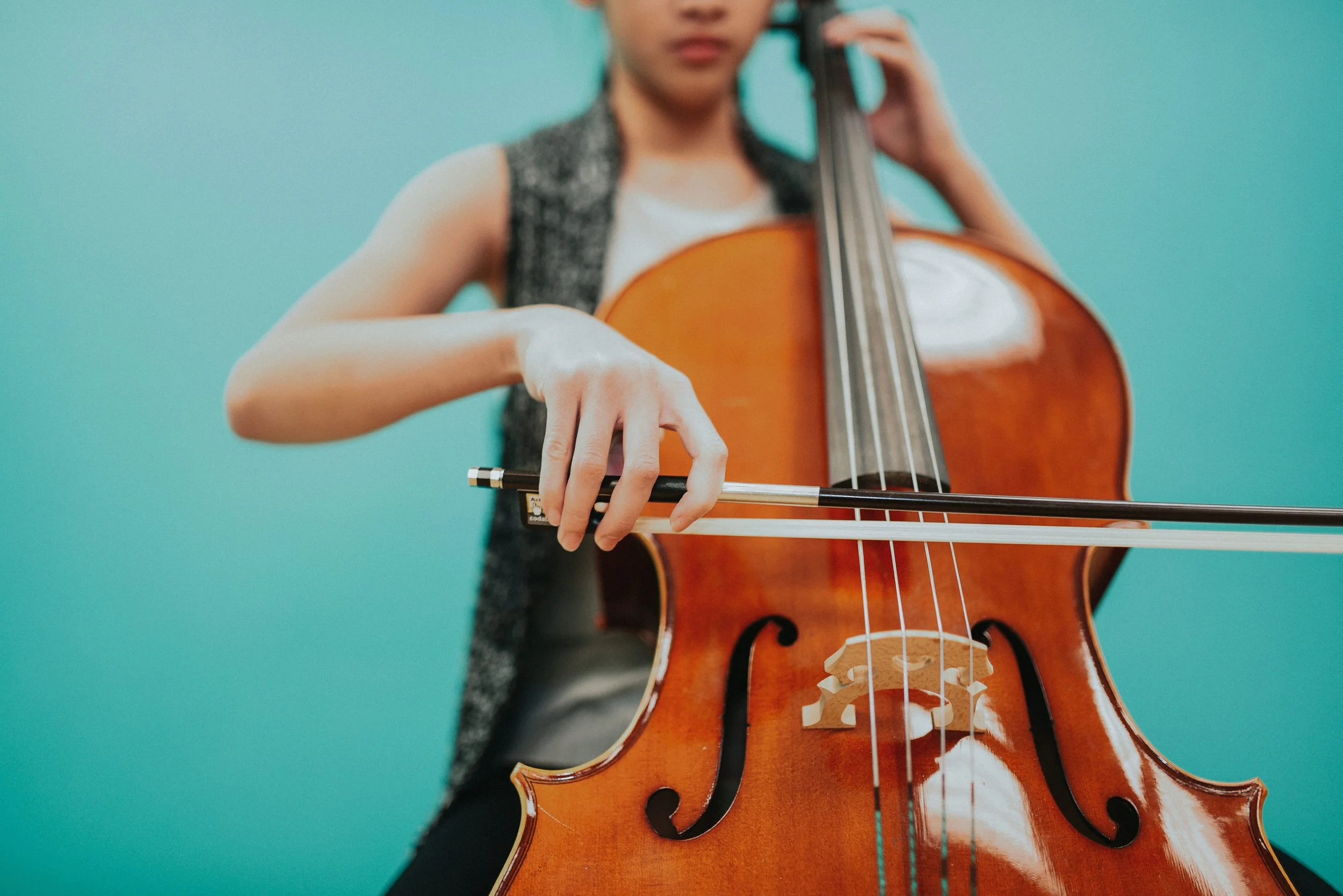 A person playing a cello, focusing on their hand holding the bow across the strings, with a teal background.