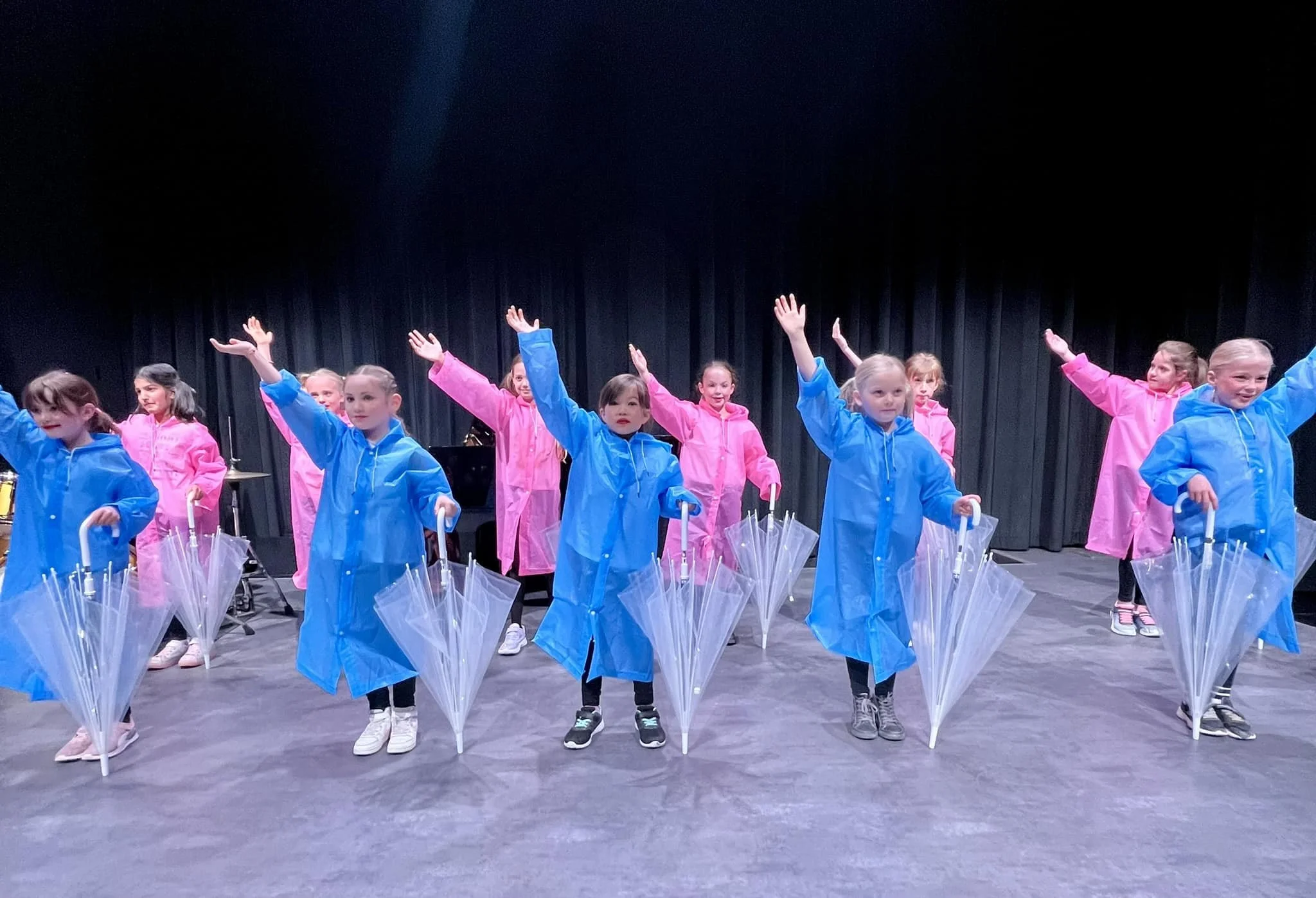 Group of young girls wearing colorful raincoats and holding transparent umbrellas, performing a dance or routine on stage with a black curtain backdrop.