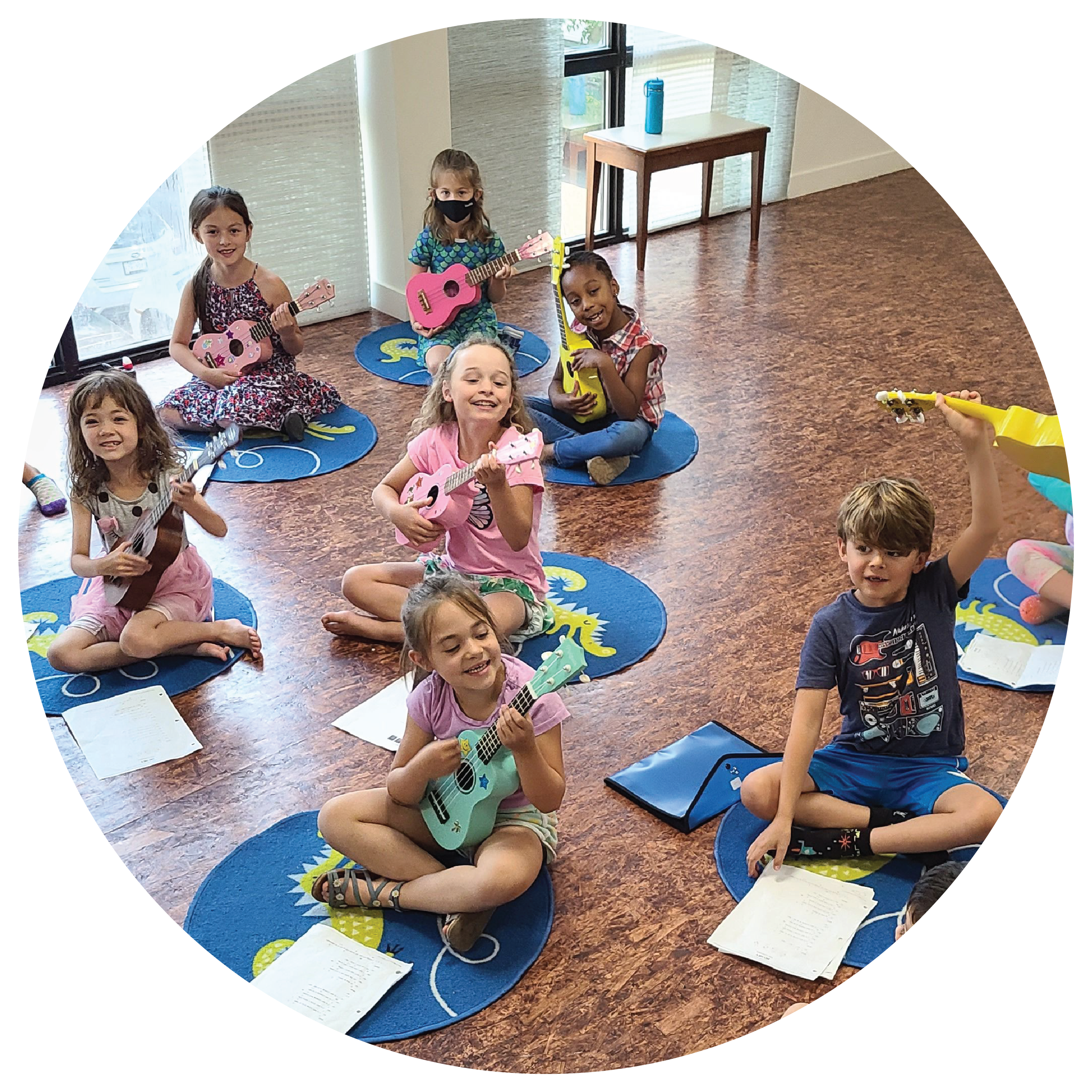 Children sitting cross-legged on blue round mats, playing small colorful guitars and singing, in a classroom with large windows and a wooden floor.