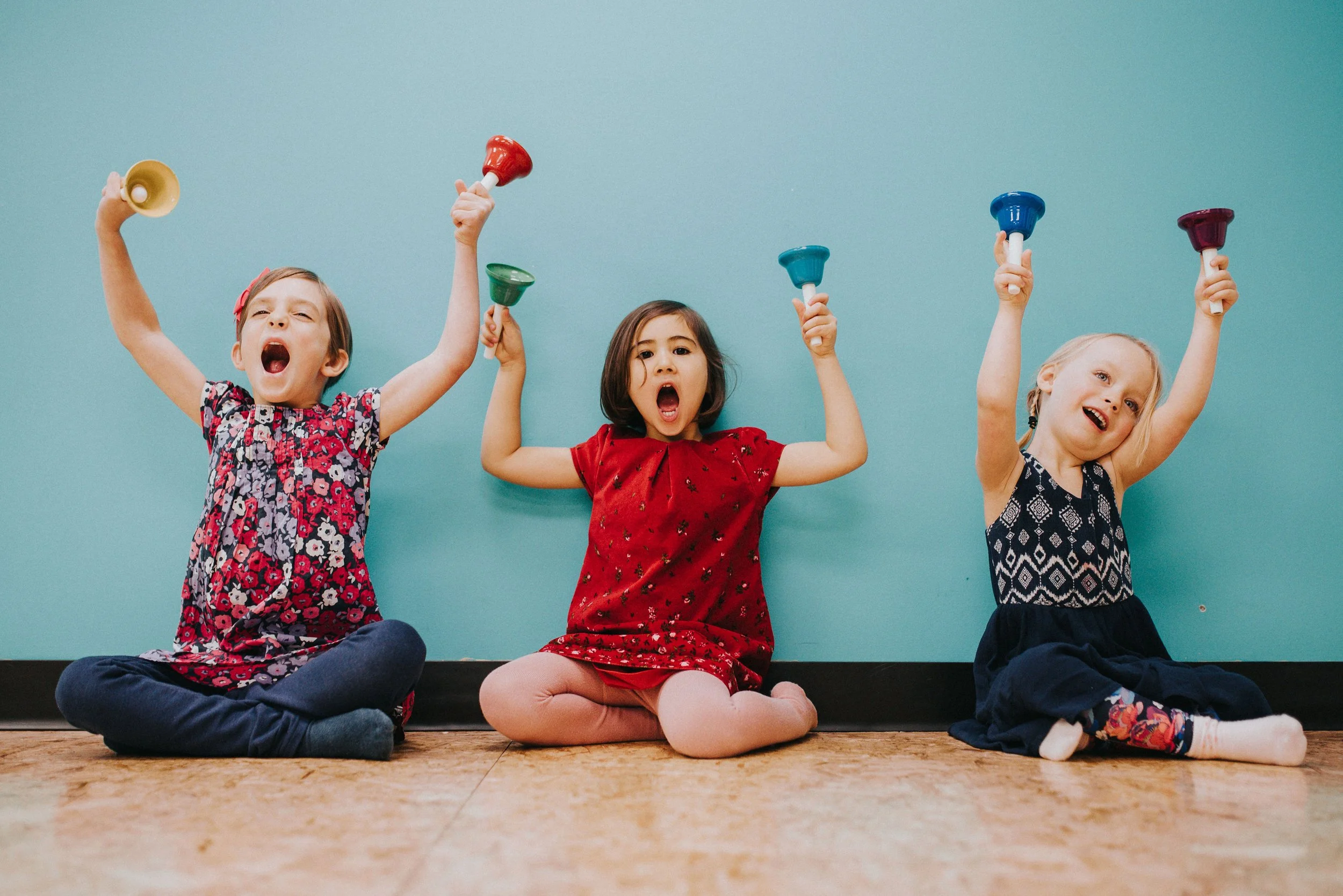 Three young girls sitting on the floor against a blue wall, raising colorful small bells in the air, with joyful expressions.