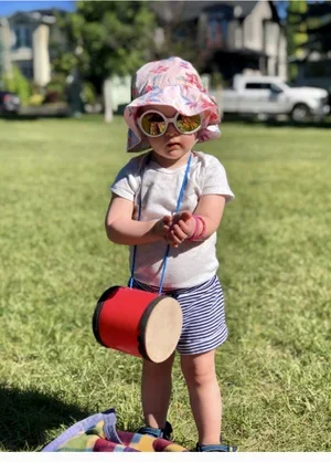 A young girl outdoors wearing a pink hat with white and pink patterns, large sunglasses, a light gray t-shirt, blue and white striped shorts, holding a small pink object, with a red and black drum hanging from her neck, standing on grass with trees and a few houses in the background.