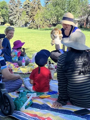 A woman holding a stuffed lion toy and entertaining a group of children sitting on a colorful picnic blanket in a park on a sunny day.