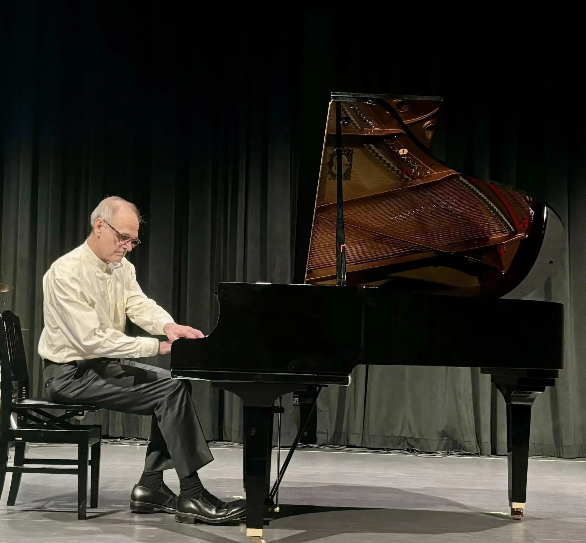 An elderly man with glasses playing the piano on a stage with black curtains.