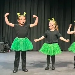 Three young girls in black t-shirts and bright green tutus performing on stage with headbands featuring antennae, dancing