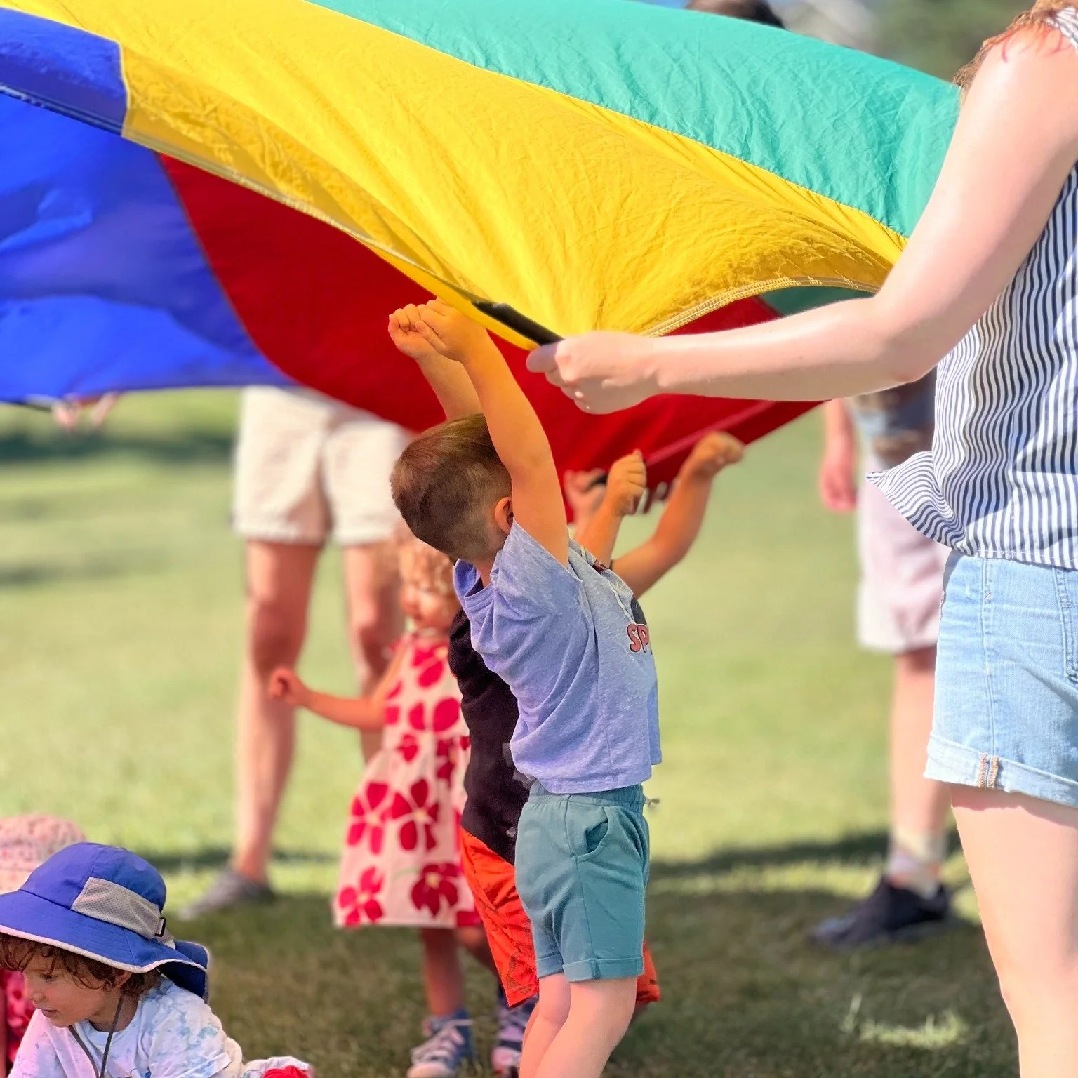 Children playing with a colorful parachute outdoors on grass, some children lifting it and others sitting underneath, with adults supervising.