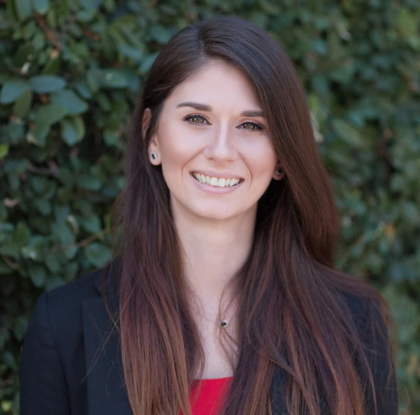 Portrait of a young woman with long brown hair, smiling, wearing a black blazer and red top, standing outdoors with green foliage background.