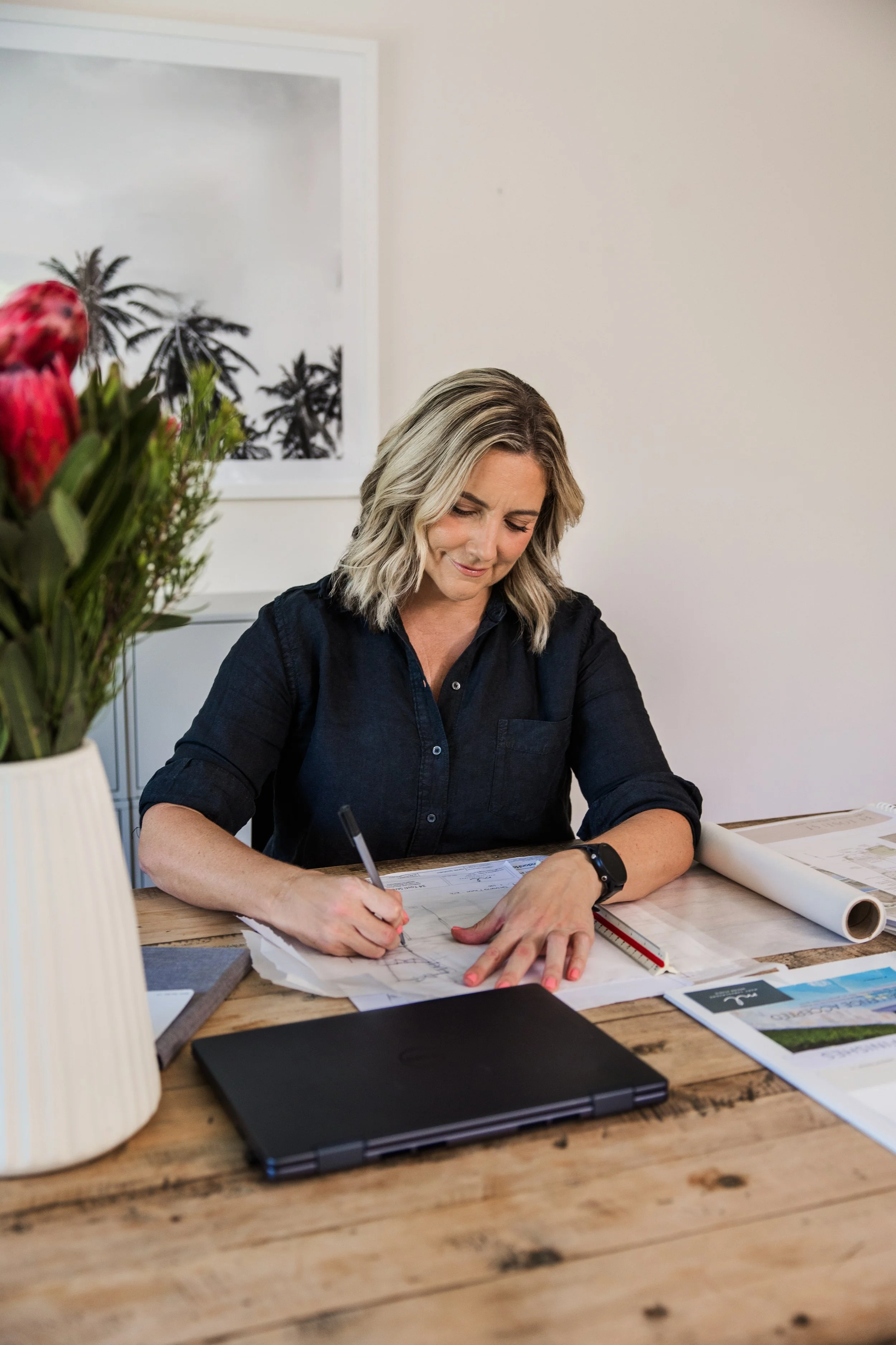 Woman with blonde hair working on architectural blueprints at a wooden table, surrounded by a laptop, rolled-up blueprints, and a blue and red ruler, with a framed picture of palm trees on the wall in the background.