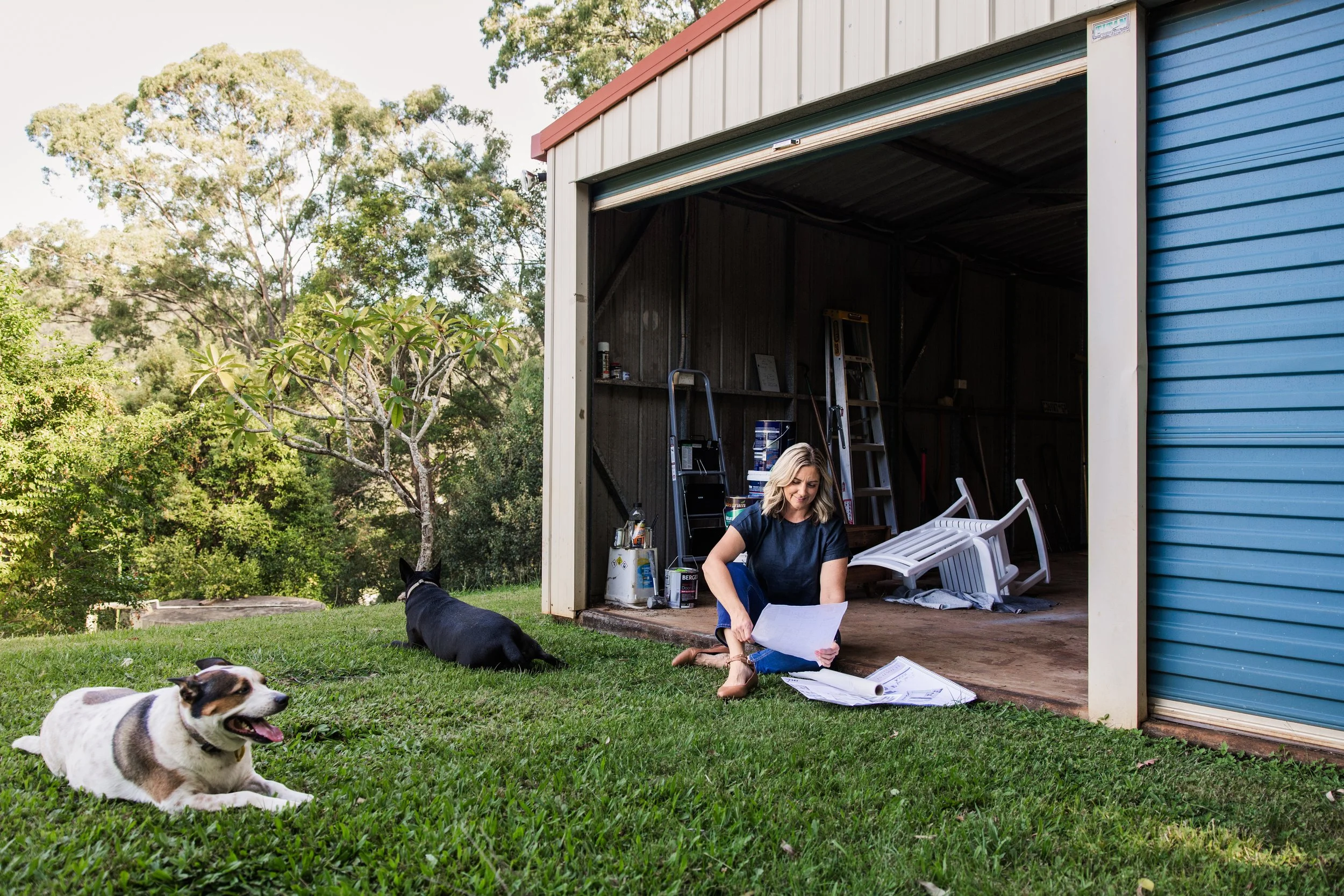 A woman sitting on the ground outside a garage, reading papers, with three dogs lying nearby. One dog is black with white markings, another is a small white dog with brown patches, and the third dog is in the garage. Trees and greenery are in the background.