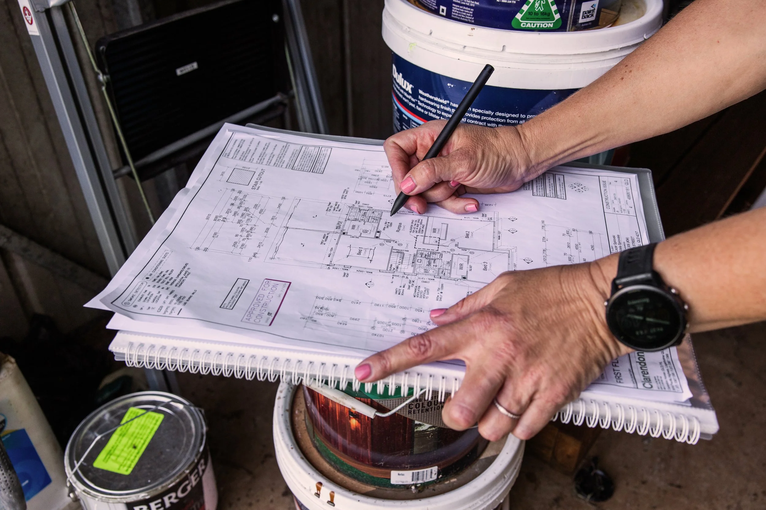 A person with a watch on their wrist holding a black pen and reviewing architectural blueprints or construction plans. The plans are placed on a spiral notebook, with several cans and buckets of paint or construction materials surrounding them.