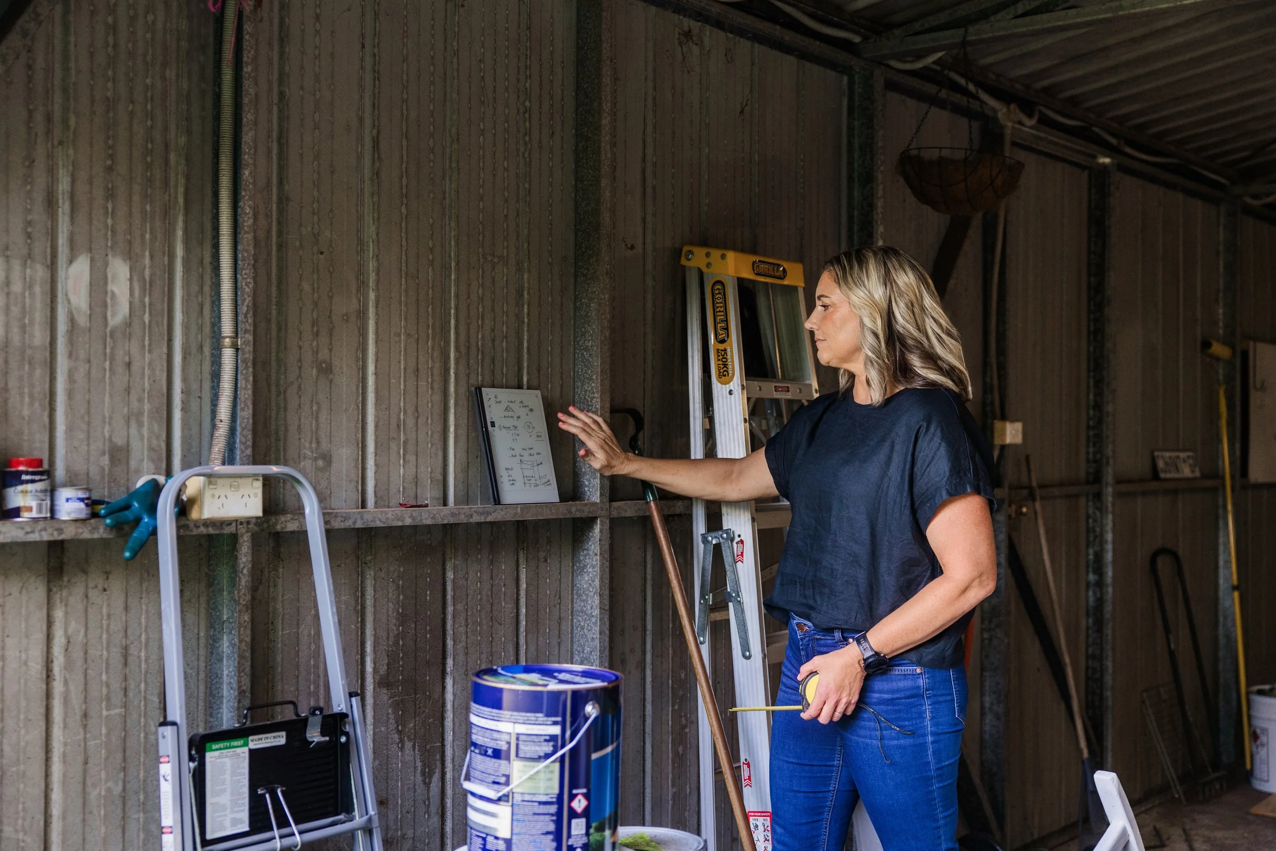 A woman with blonde hair wearing a black T-shirt and jeans stands in a garage or workshop, facing a wall with various tools and equipment. She is holding a tool in her right hand and appears to be working or inspecting something. There is a ladder, paint cans, and other workshop items visible around her.