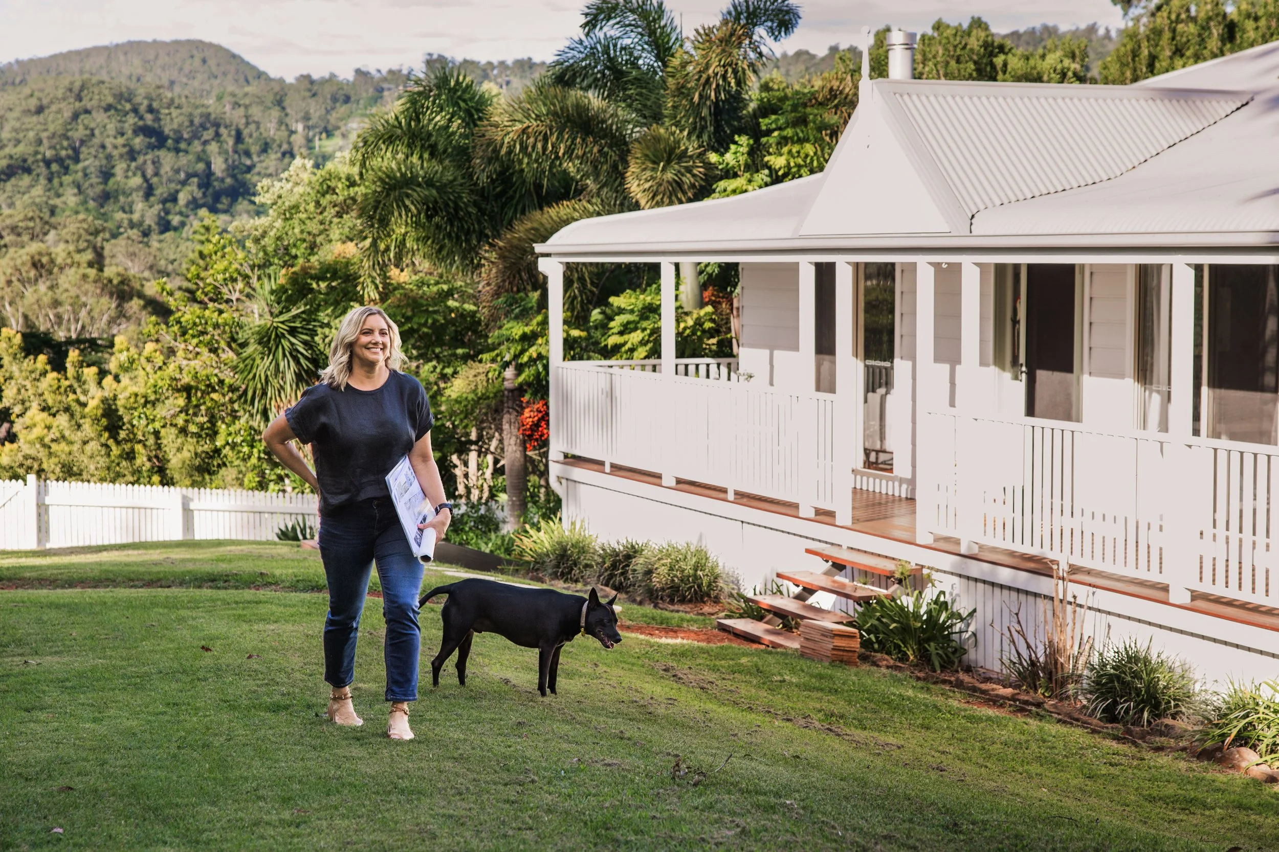 A woman smiling and holding a rolled-up document walking in a lush green backyard with a black dog nearby. A white house with a porch and multiple windows is visible, surrounded by trees and mountains in the background.