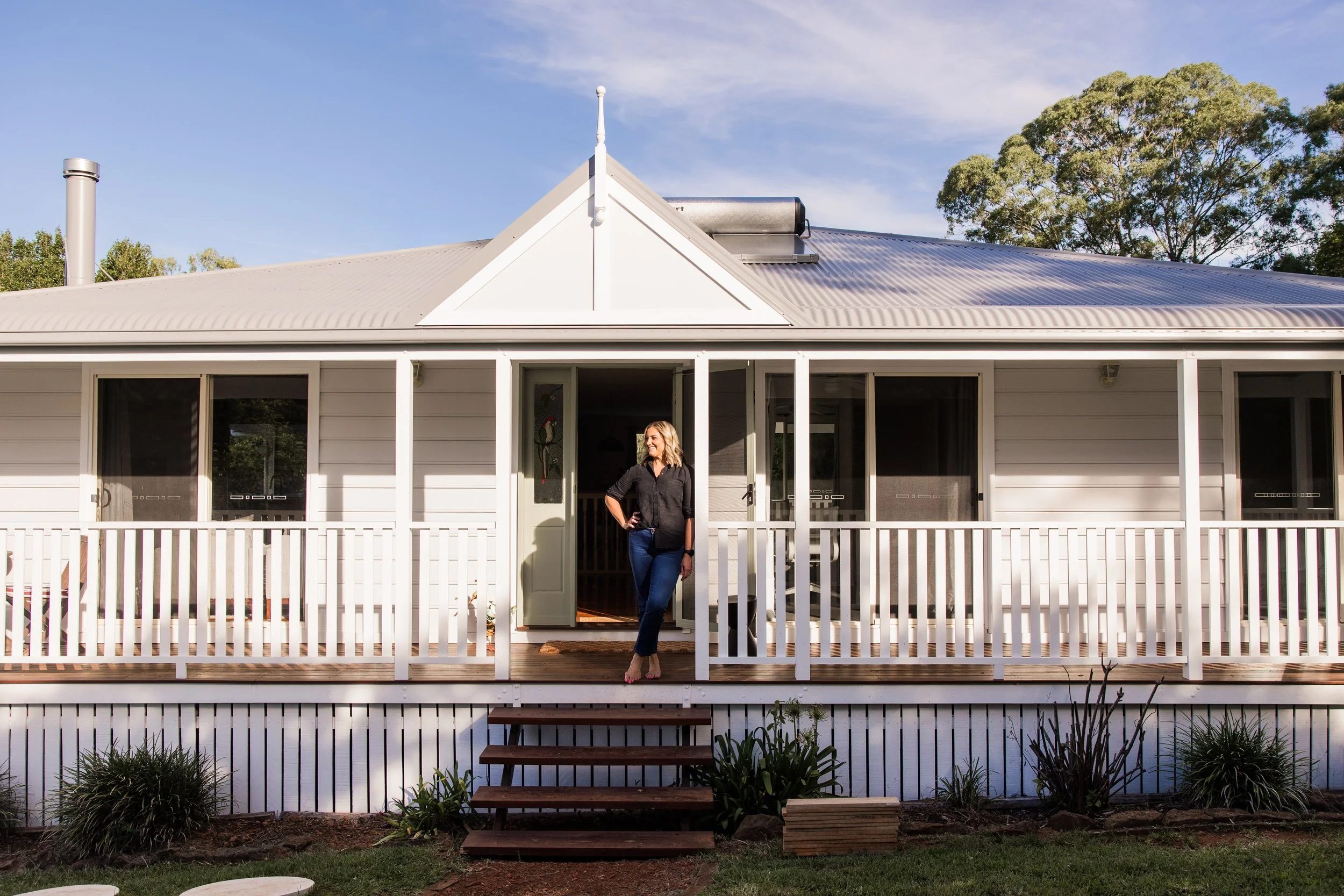 Woman standing on porch of white house with picket fence, in front of stairs, with garden plants at the bottom.