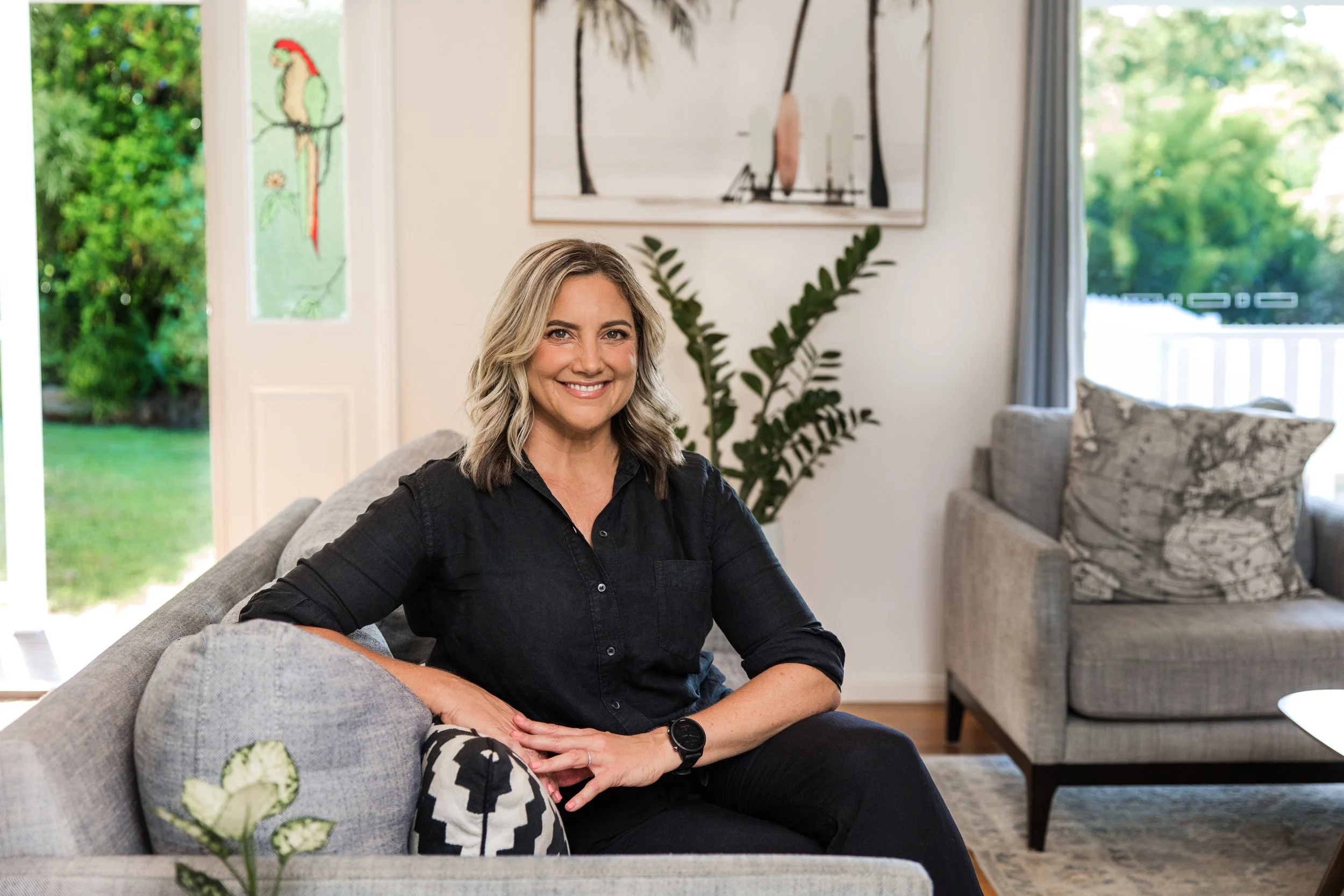 A smiling woman with shoulder-length blonde hair sitting on a gray sofa in a living room, with greenery visible outside the open door behind her, a large artwork with palm trees hanging on the wall, and a tall plant beside her.