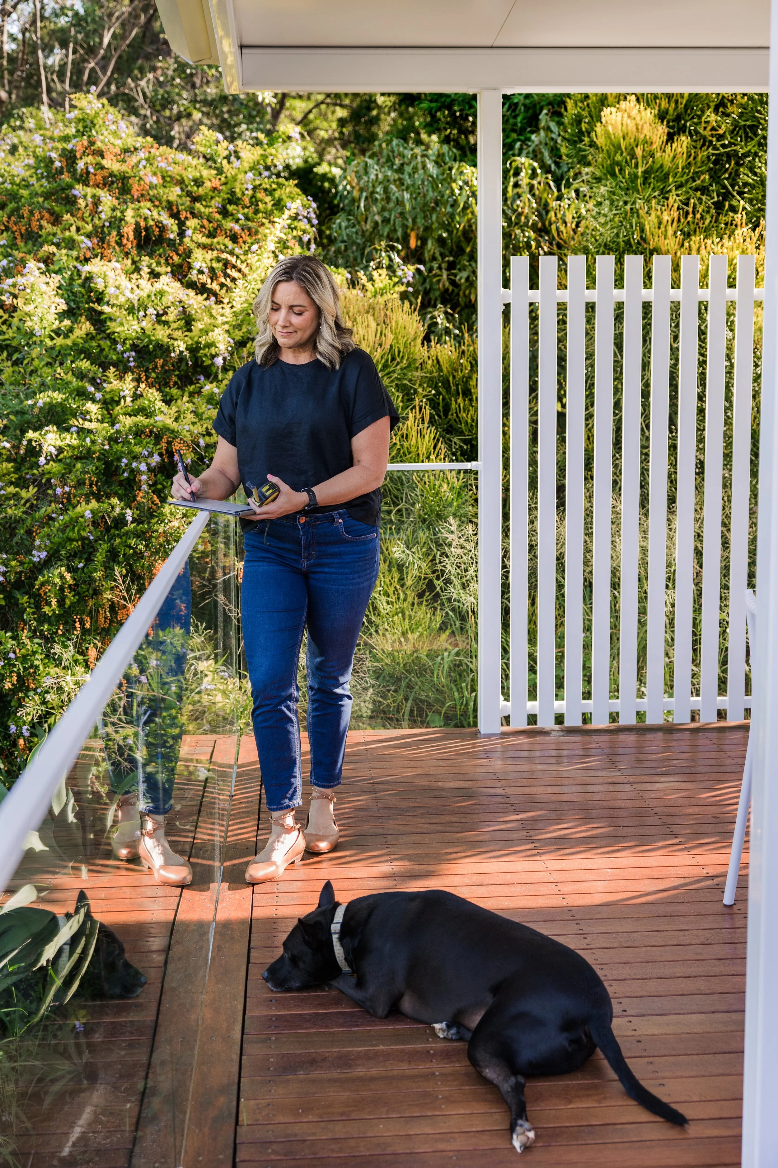 A woman with shoulder-length blonde hair wearing a black t-shirt, blue jeans, and beige shoes, standing on a wooden deck outdoors. She is holding a pen and a notepad, with a black dog lying on the deck nearby. The background features lush green plants and a white fence.
