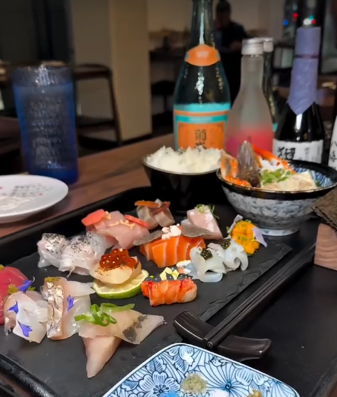 Assorted sushi and sashimi on a black platter, with bowls of rice, soy sauce, and bottles of drinks in the background.
