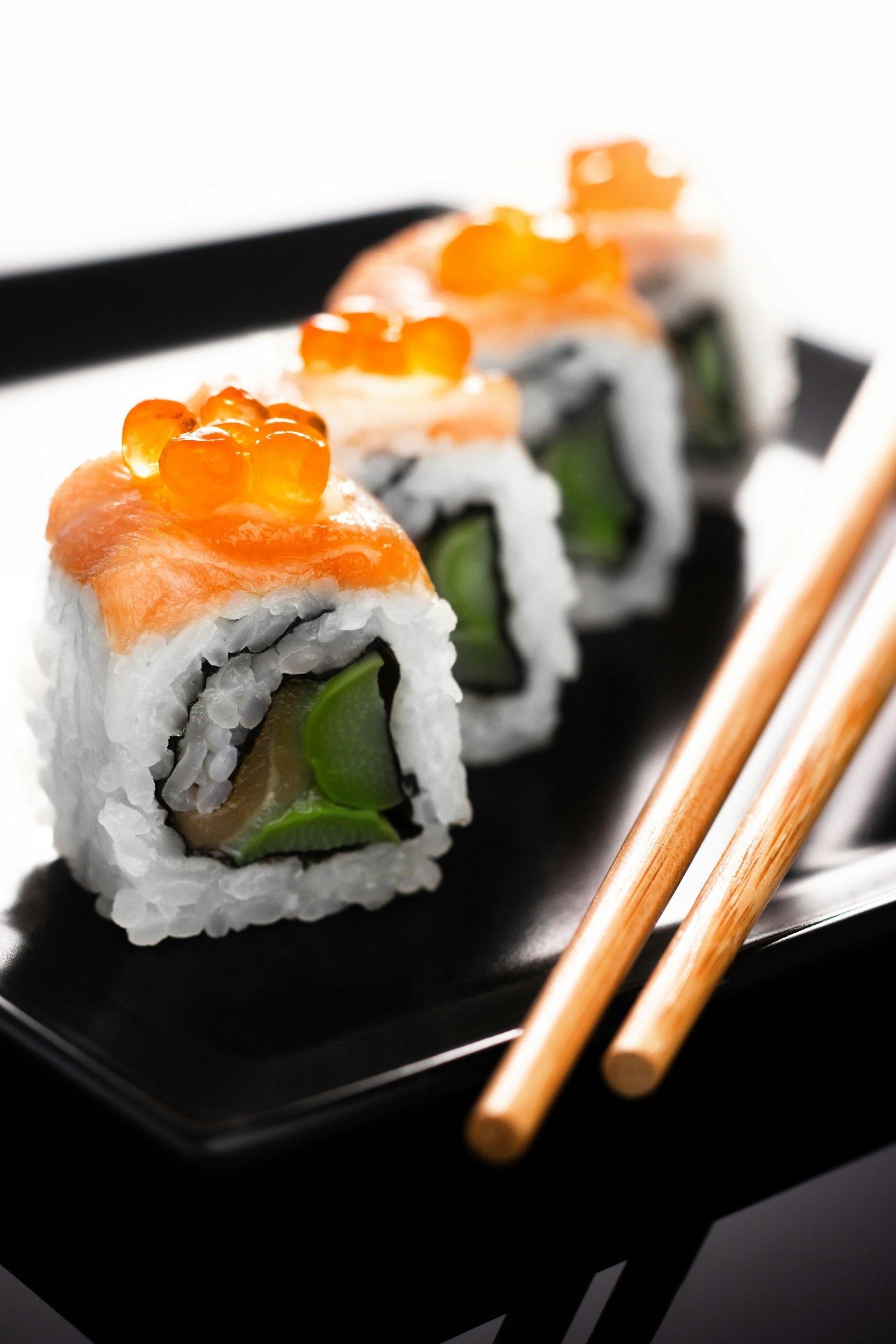 Close-up of three sushi rolls topped with salmon and orange fish roe on a black plate with chopsticks.