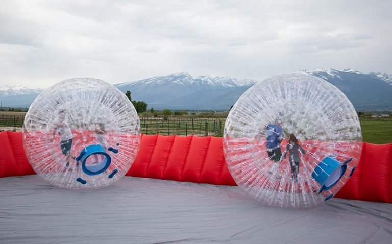 Two people in inflatable bumper balls on an outdoor inflatable arena with mountains and cloudy sky in the background.